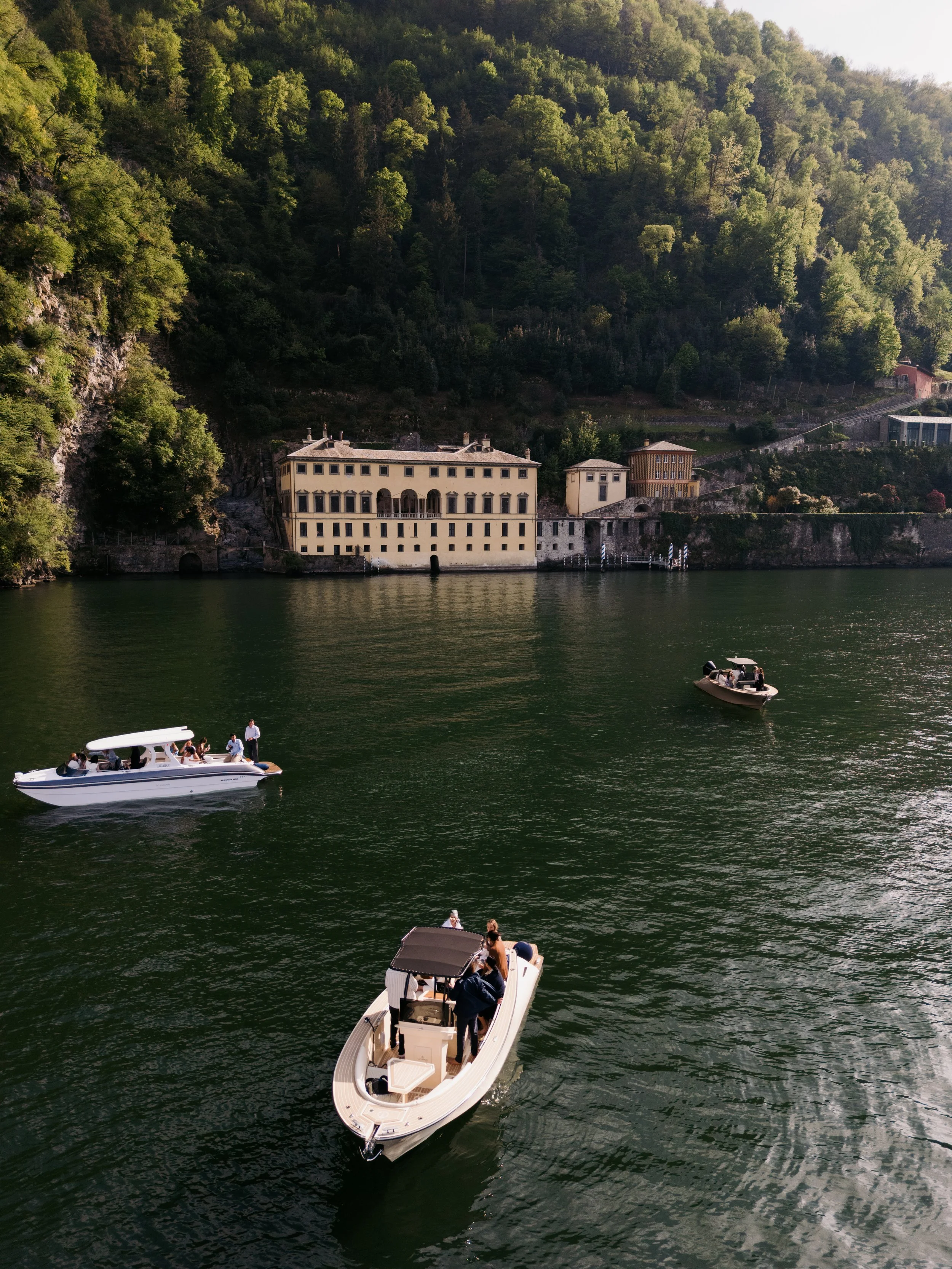 People boating on a lake with a hillside covered in trees and historic buildings in the background.