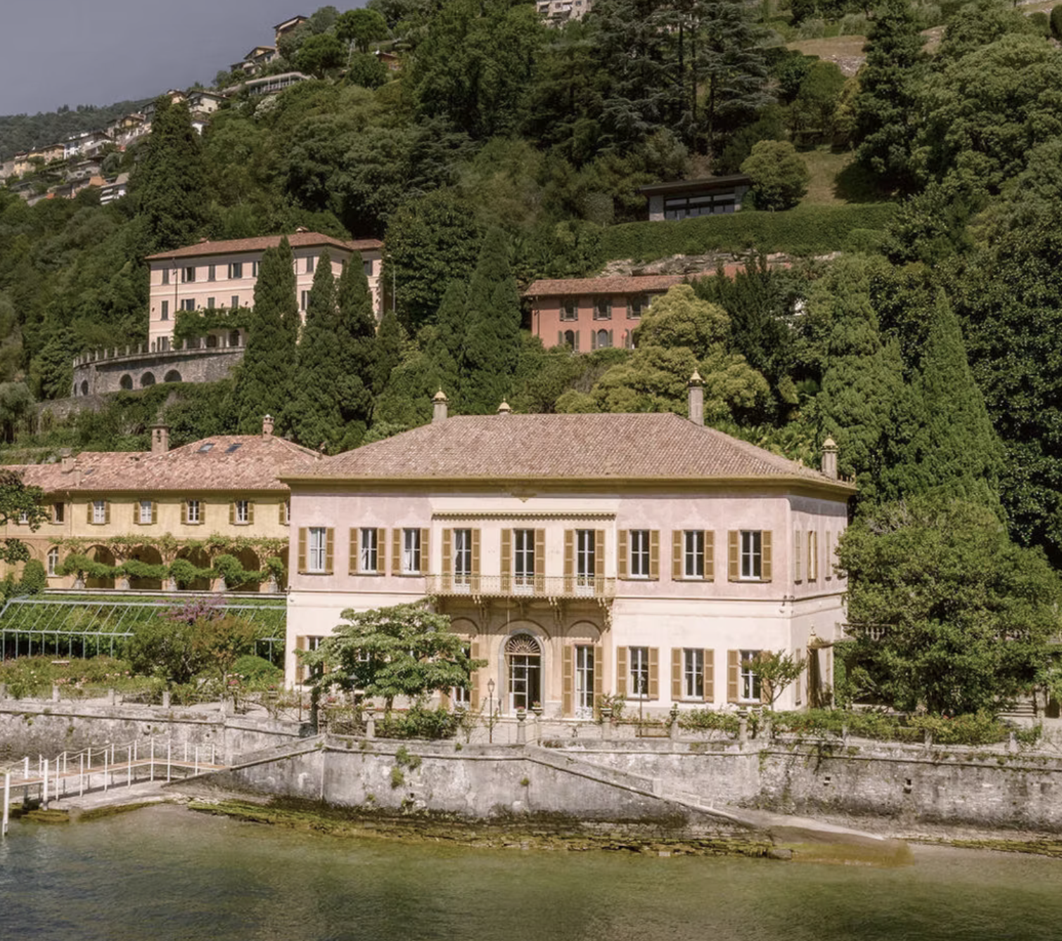 Lake Como villa with a tiled roof situated by a river, with greenery and hills in the background.