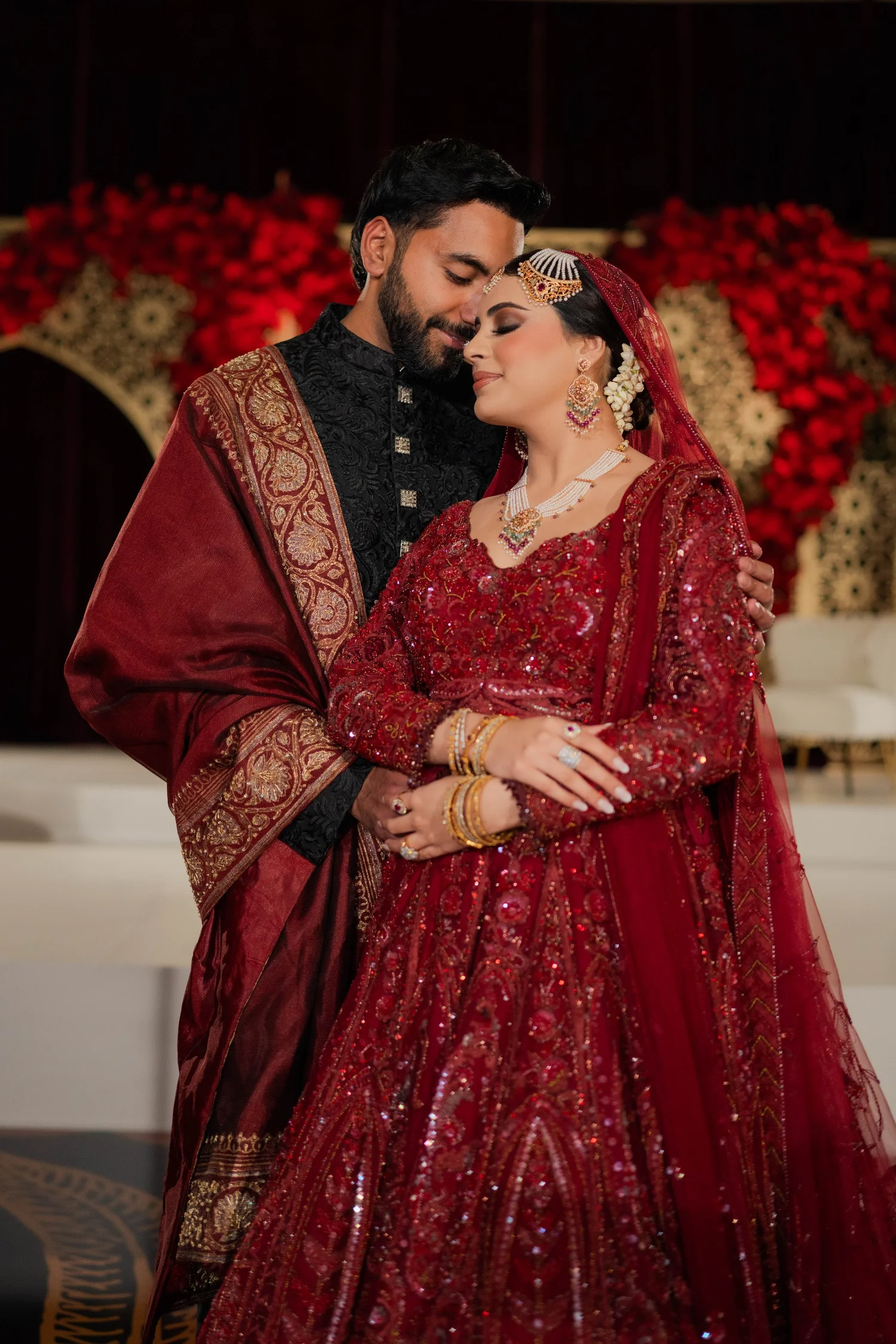 A bride and groom in traditional red South Asian wedding attire, in front of red florals and a gold arch