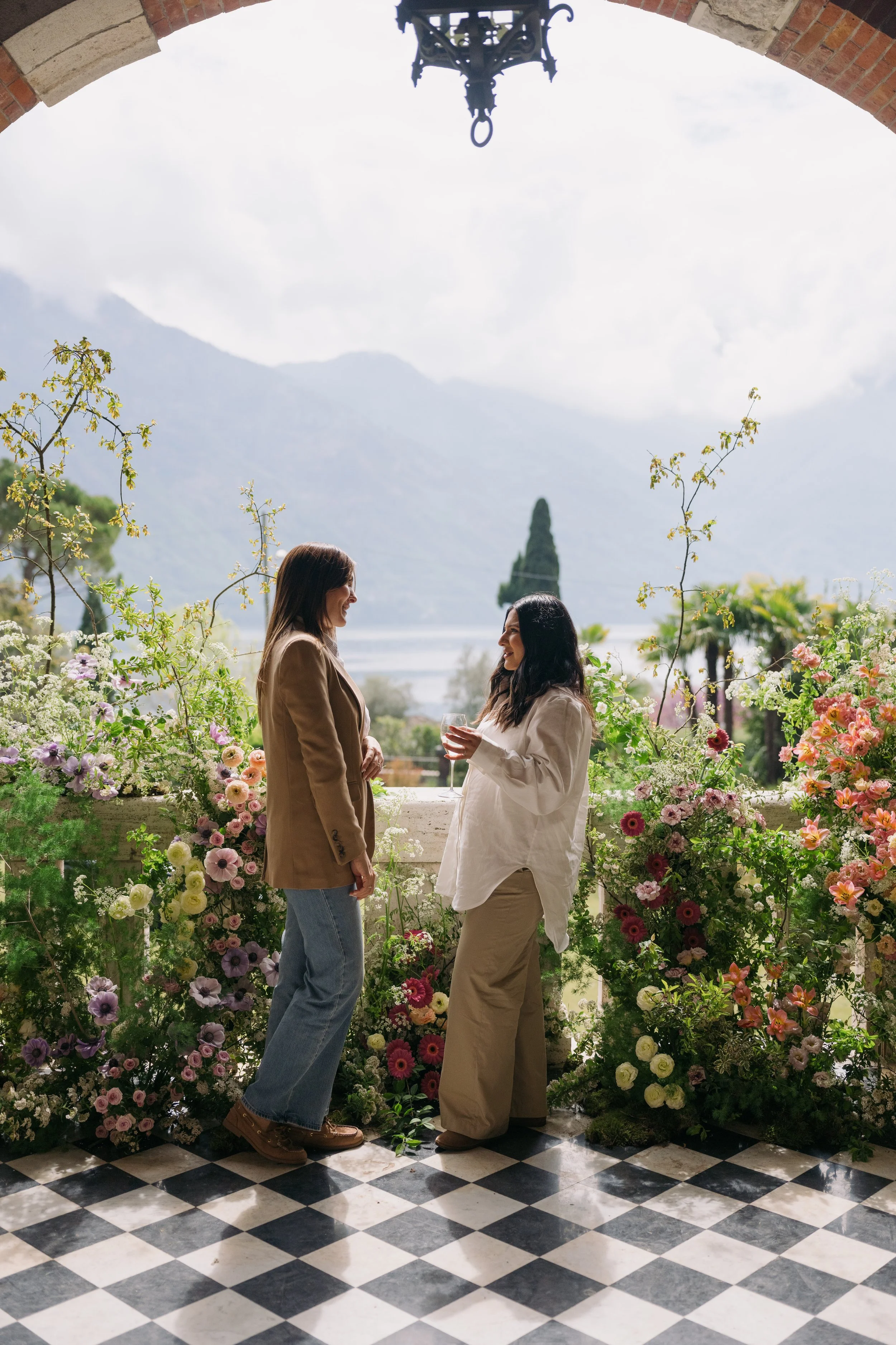 Wedding Planner in Lake Como meeting with client and venue surrounded by an abundance of colorful flowers, smiling and facing each other, with a lake and mountains in the background under a cloudy sky.