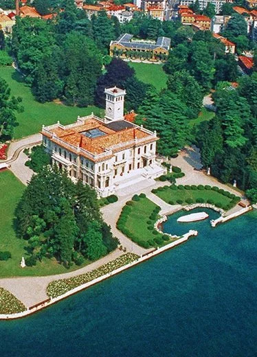 Villa Serbelloni Bellagio Wedding Planner at the mansion with a red tiled roof, situated on a lakeside with a landscaped garden, fountain, and walking paths, surrounded by lush green trees.