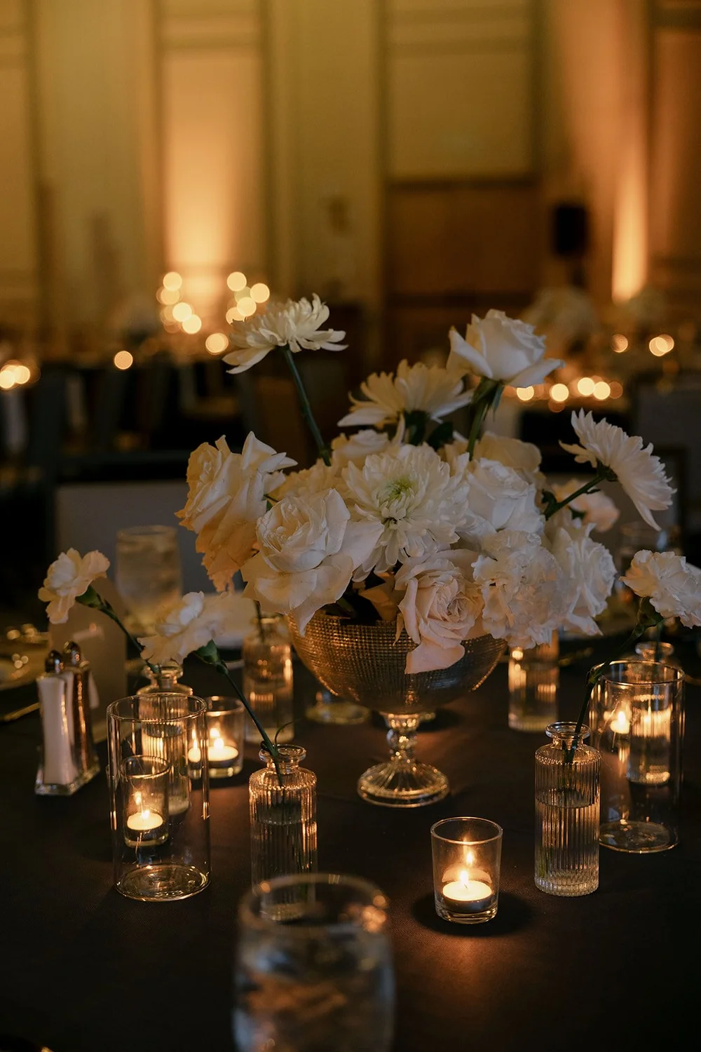 A centerpiece of white flowers in a silver vase surrounded by candles