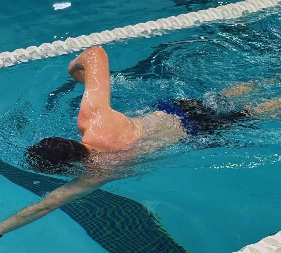 Swimmer in a pool, reaching for the pool bottom, wearing dark swimming trunks.