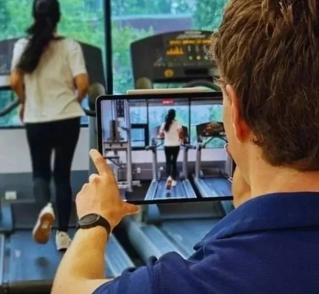 Man taking a photo of a woman walking on a treadmill at a gym with a tablet.