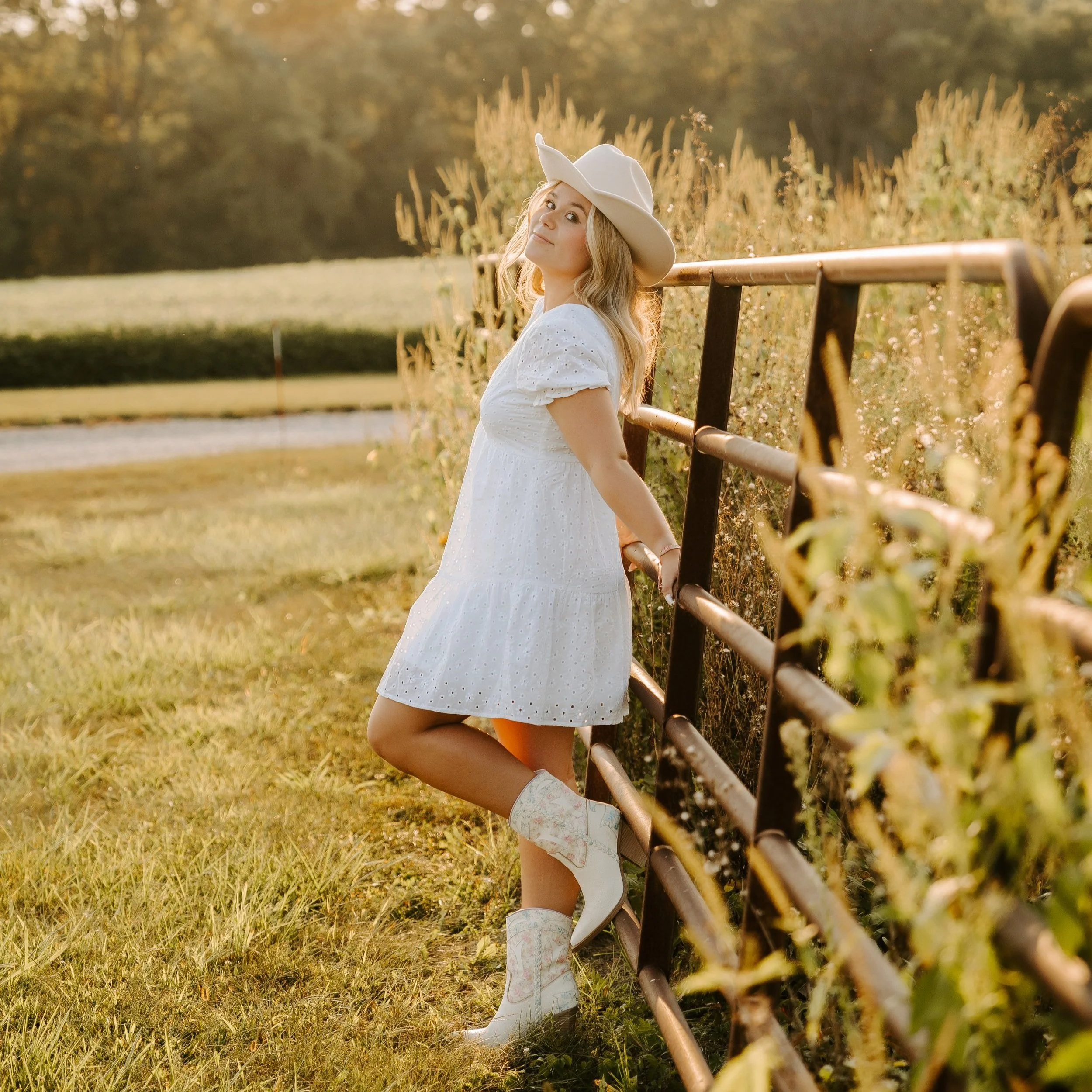 Girl in a white dress and cowboy boots leaning against a fence in a grassy field at sunset.