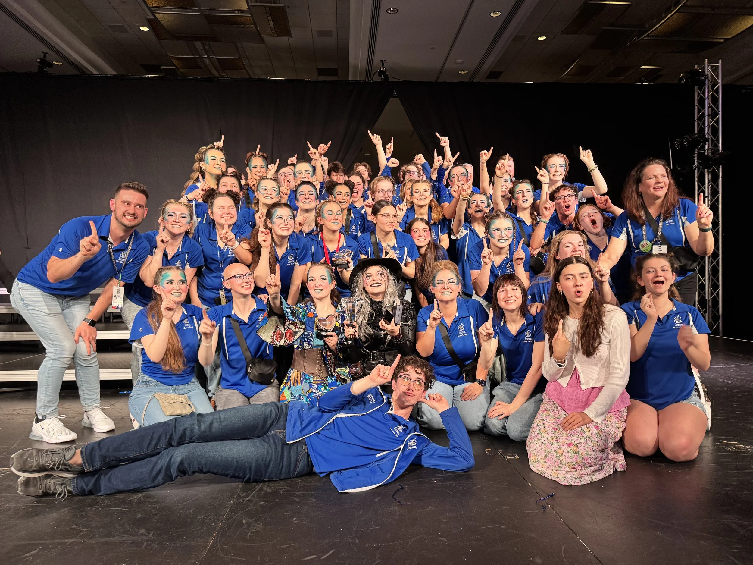 A large group of smiling young people and a few adults, all wearing blue shirts, posing on stage with some making peace signs and pointing upwards. The group appears to be celebrating or attending an event.