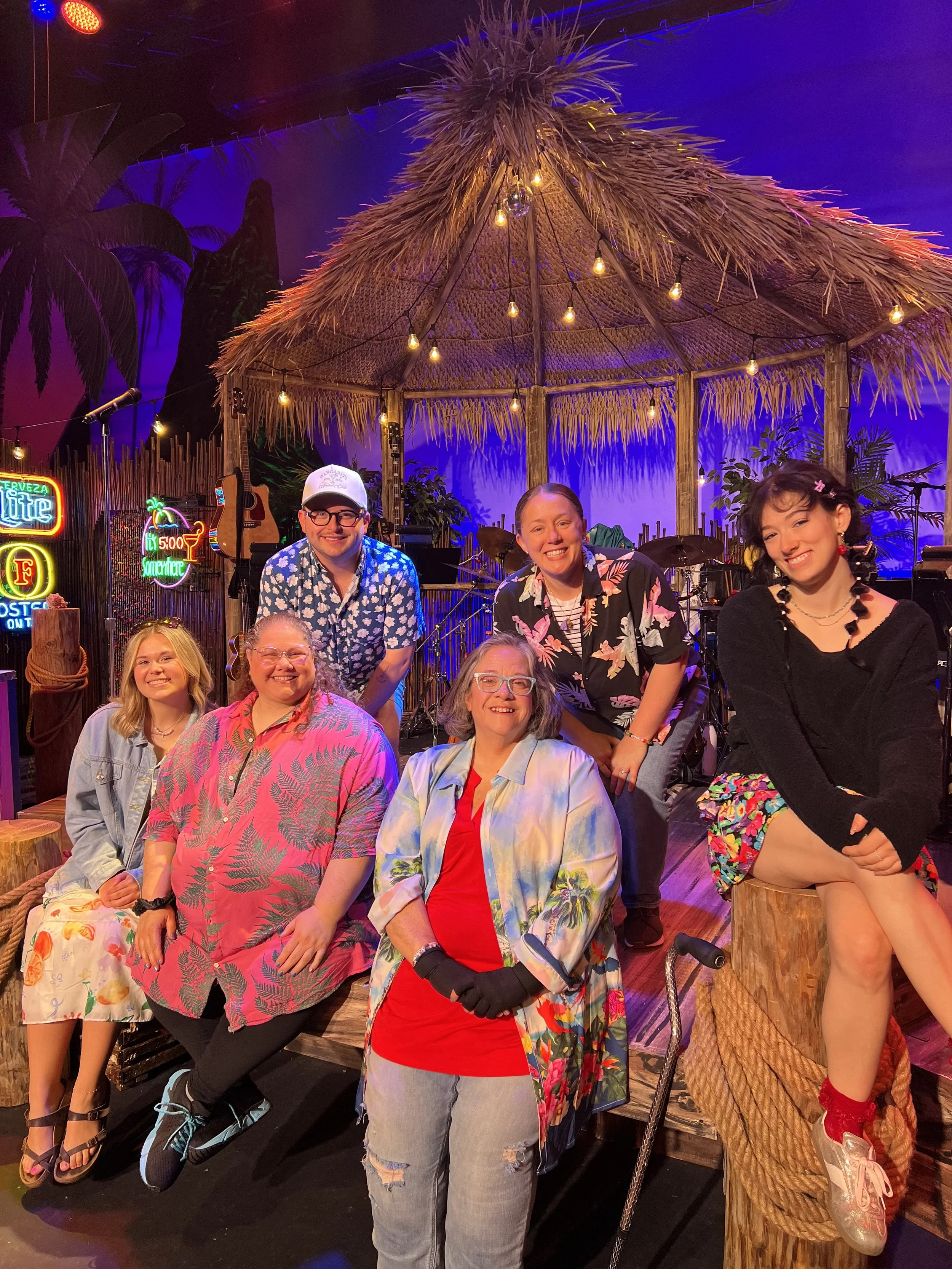 Group of six people posing under a tiki hut with tropical decorations and colorful neon signs in a lively, tropical-themed setting at the Fort Wayne Civic Theatre's production of Escape to Margaritaville.