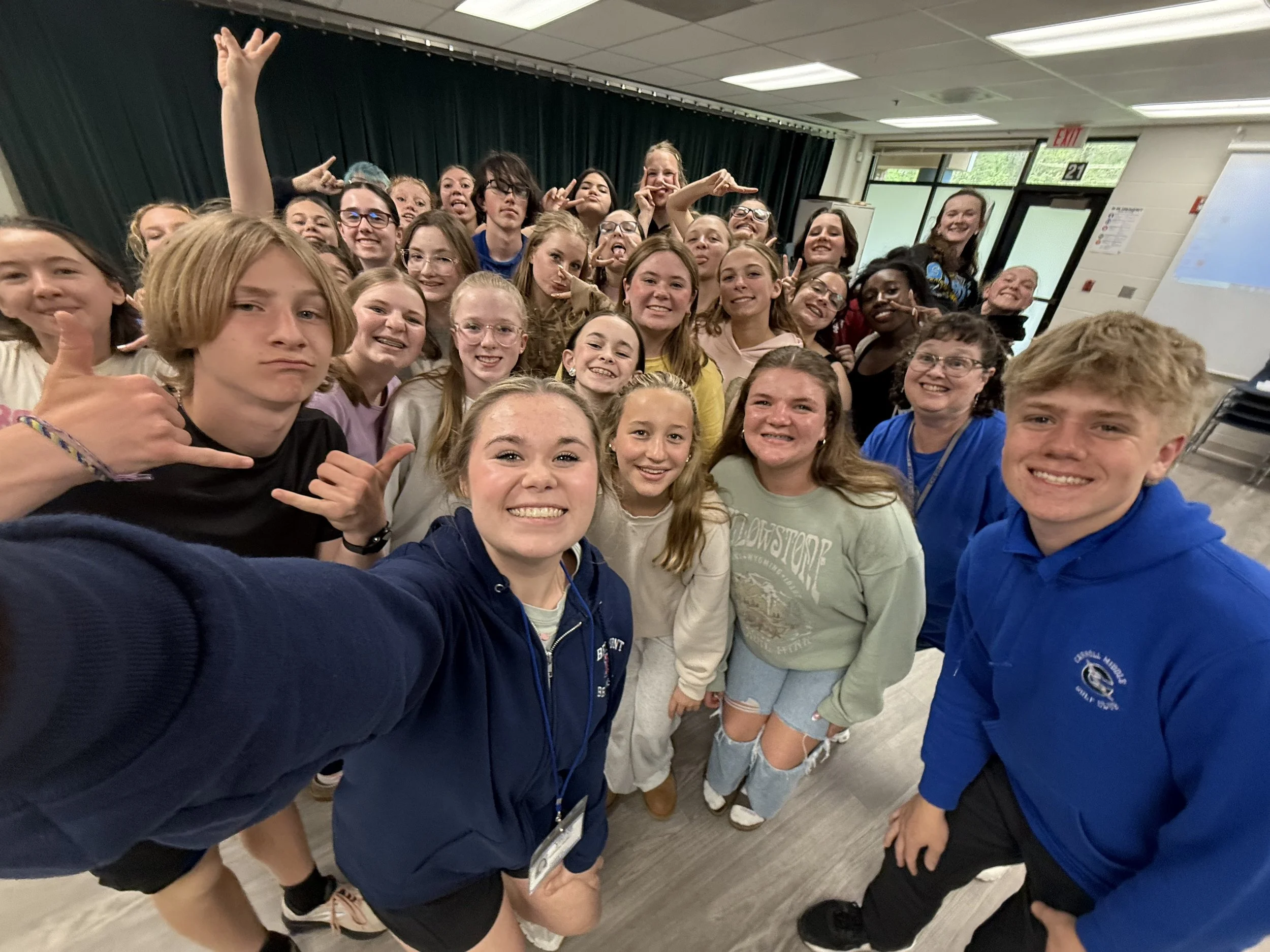 Group of students and teachers taking a selfie in a classroom, smiling and making various gestures.