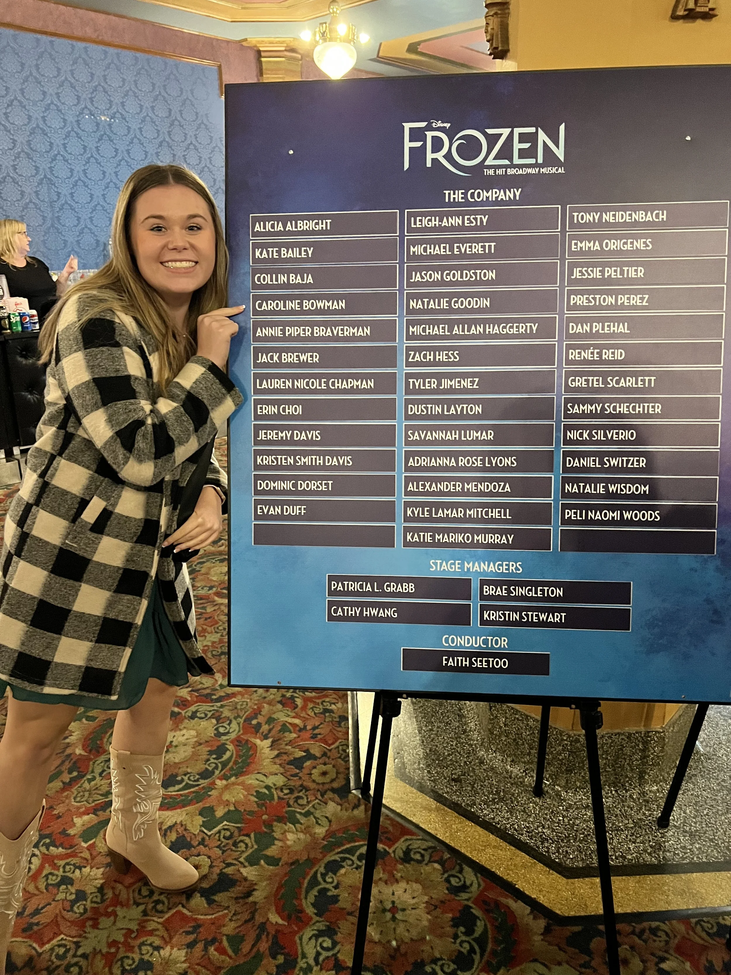 Isabella smiling and pointing at a large board displaying cast and crew members for the musical 'Frozen' at a promotional event, with pink, blue, and gold decorative indoor setting.