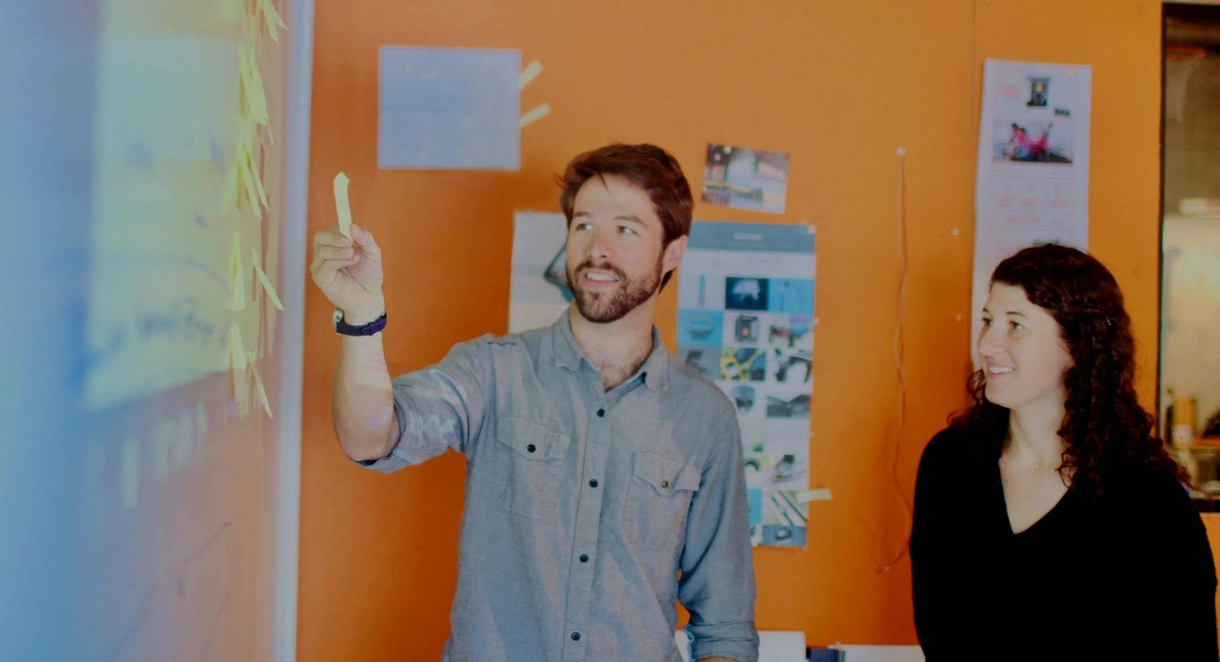 Two people, a man and a woman, in an office setting. The man is writing on a whiteboard with a marker, and the woman is smiling, watching him. The background features a bright orange wall with various pictures and papers displayed.