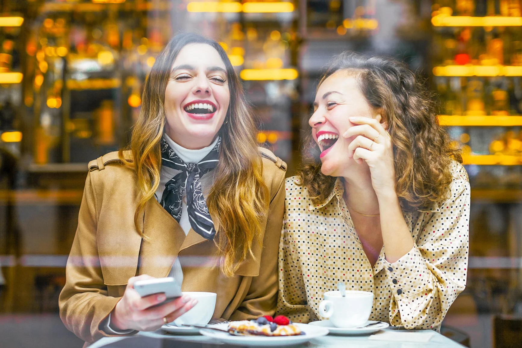 Two women laughing and enjoying coffee at a cafe with a dessert and berries on the table.