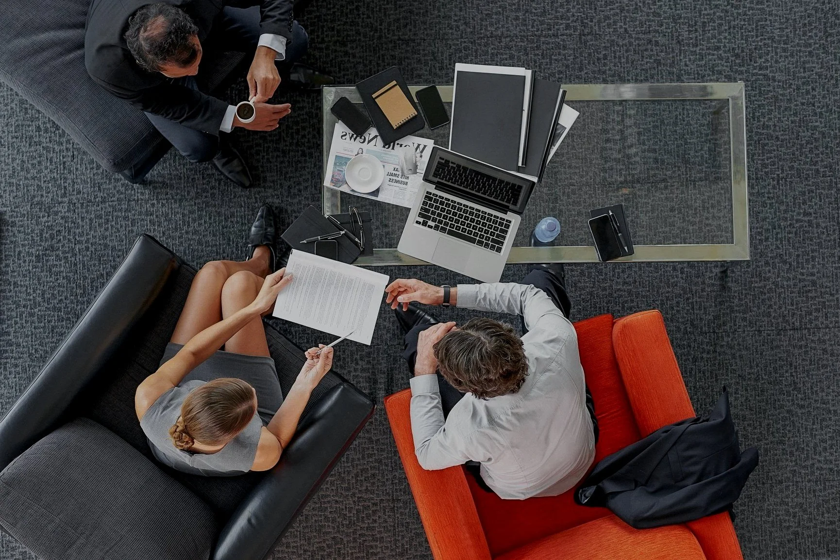Top-down view of three professionals in a business meeting in an office, seated around a glass table with laptops, notebooks, documents and coffee cups.
