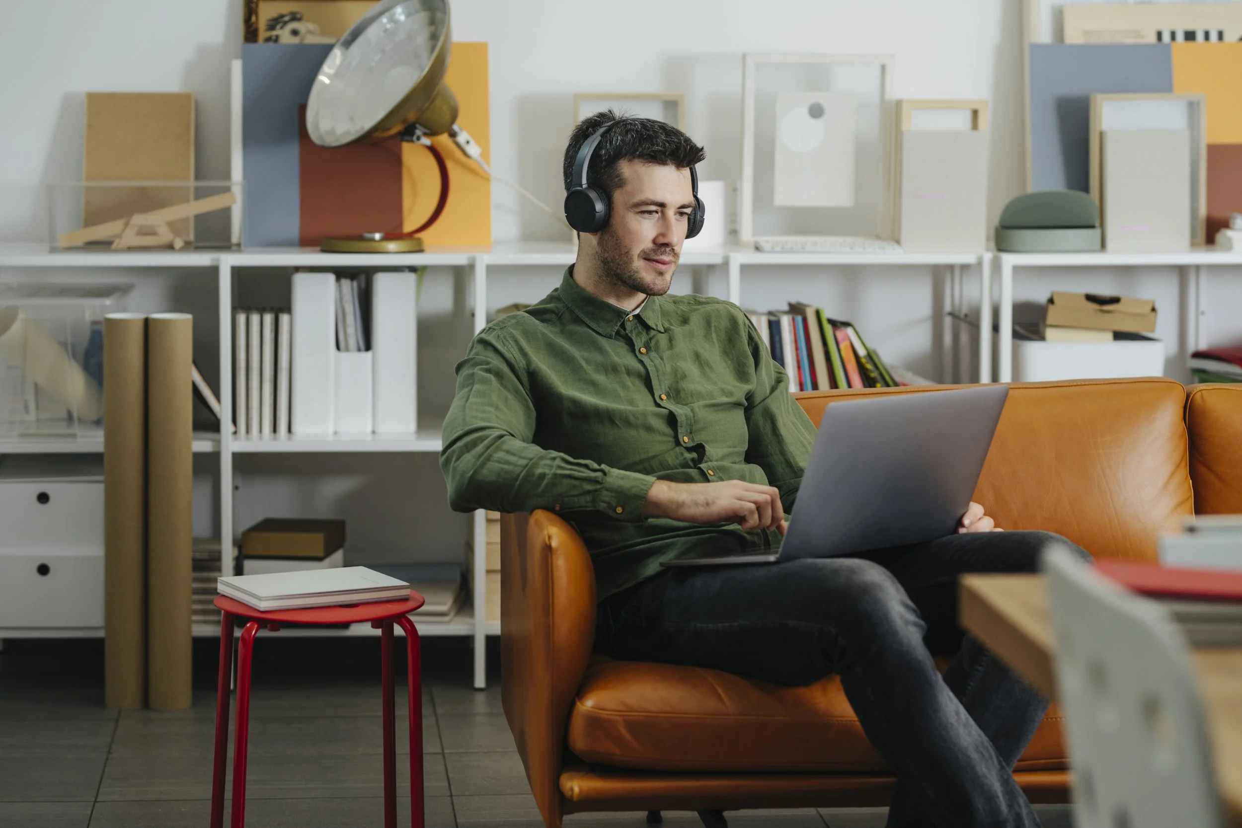 A man sitting on a tan leather couch with a laptop, wearing headphones, in a modern room with shelves of books and decorative items.
