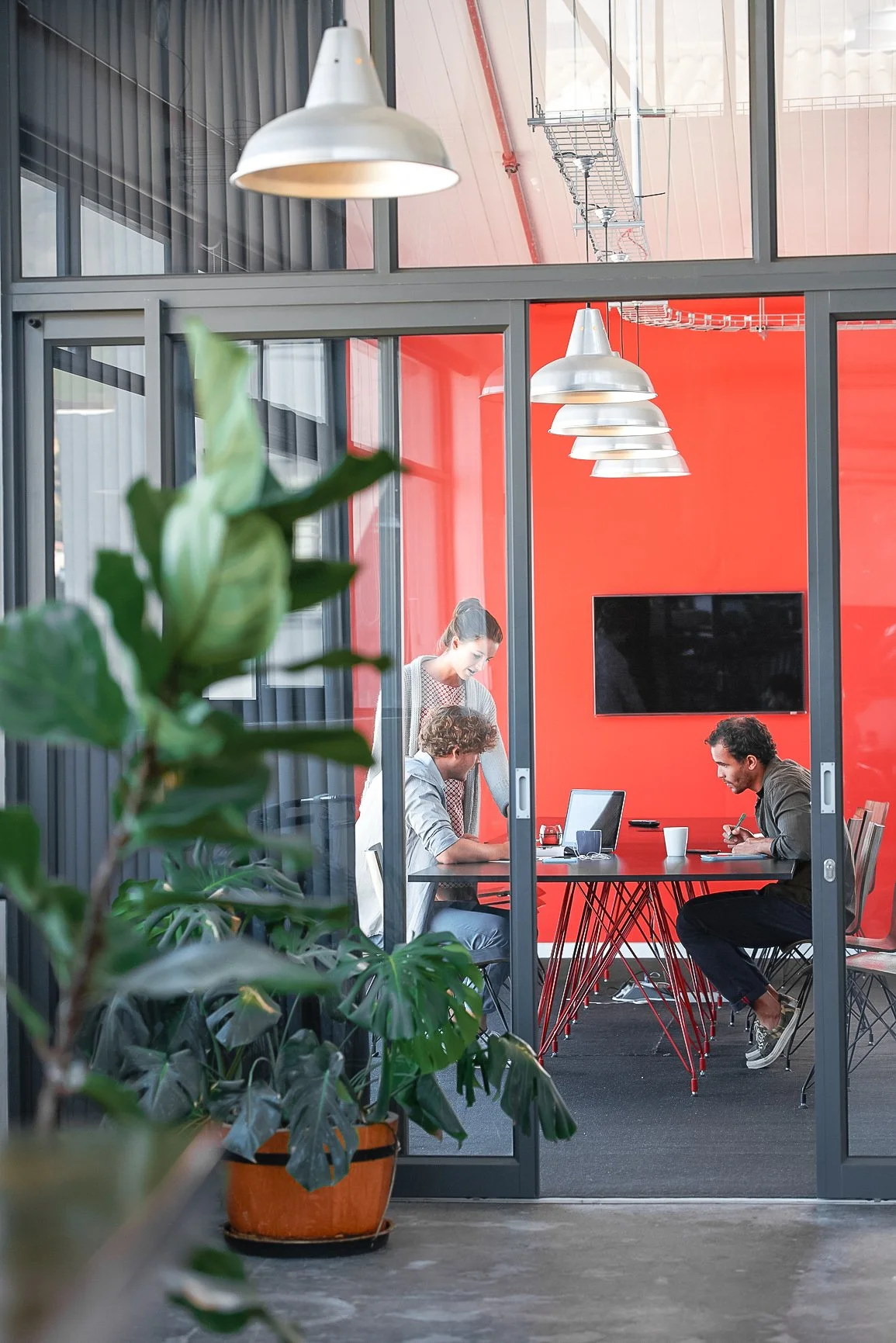 Three people in a meeting room, two seated at a red table with laptops and one standing, with a large TV on the red wall, all viewed through a glass door with a plant in the foreground.