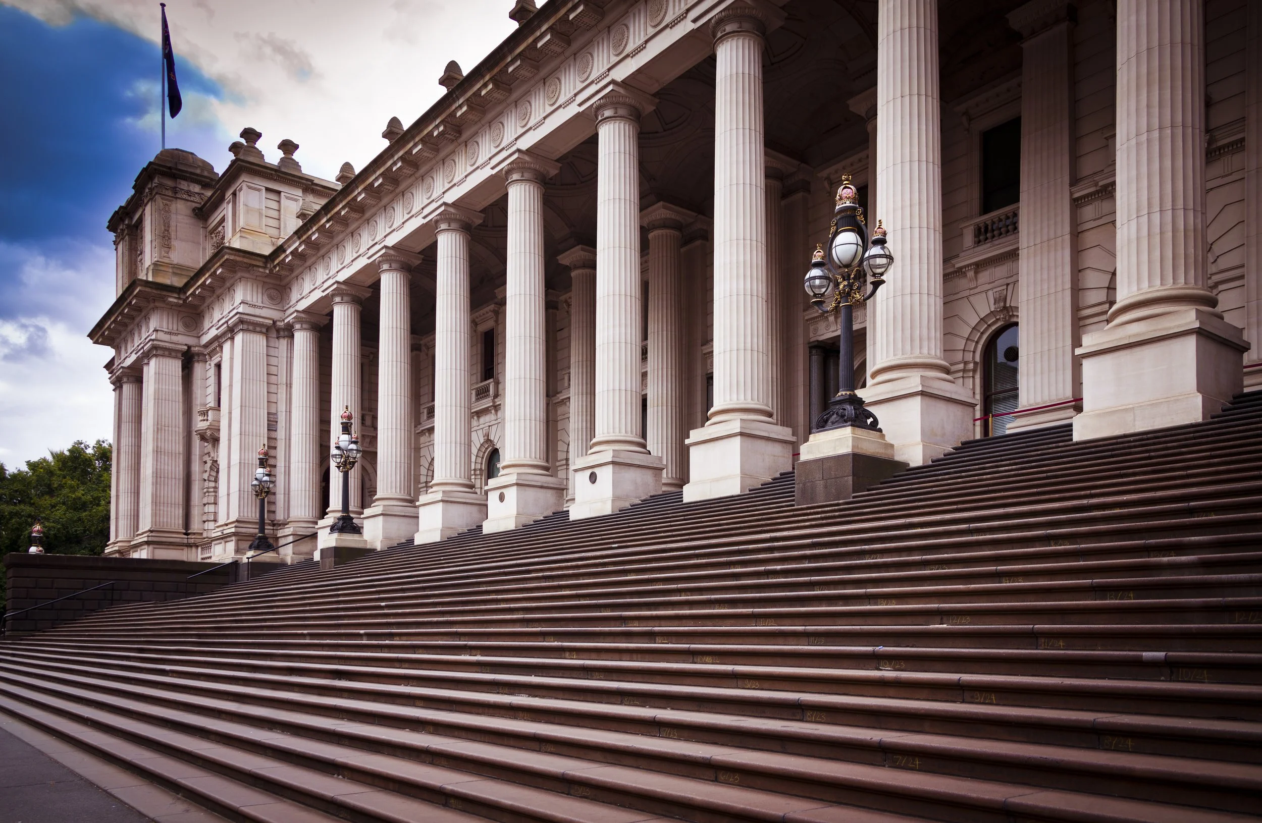 Front view of a grand neoclassical building with large steps, tall columns, ornate lamp posts, and flags in the background.