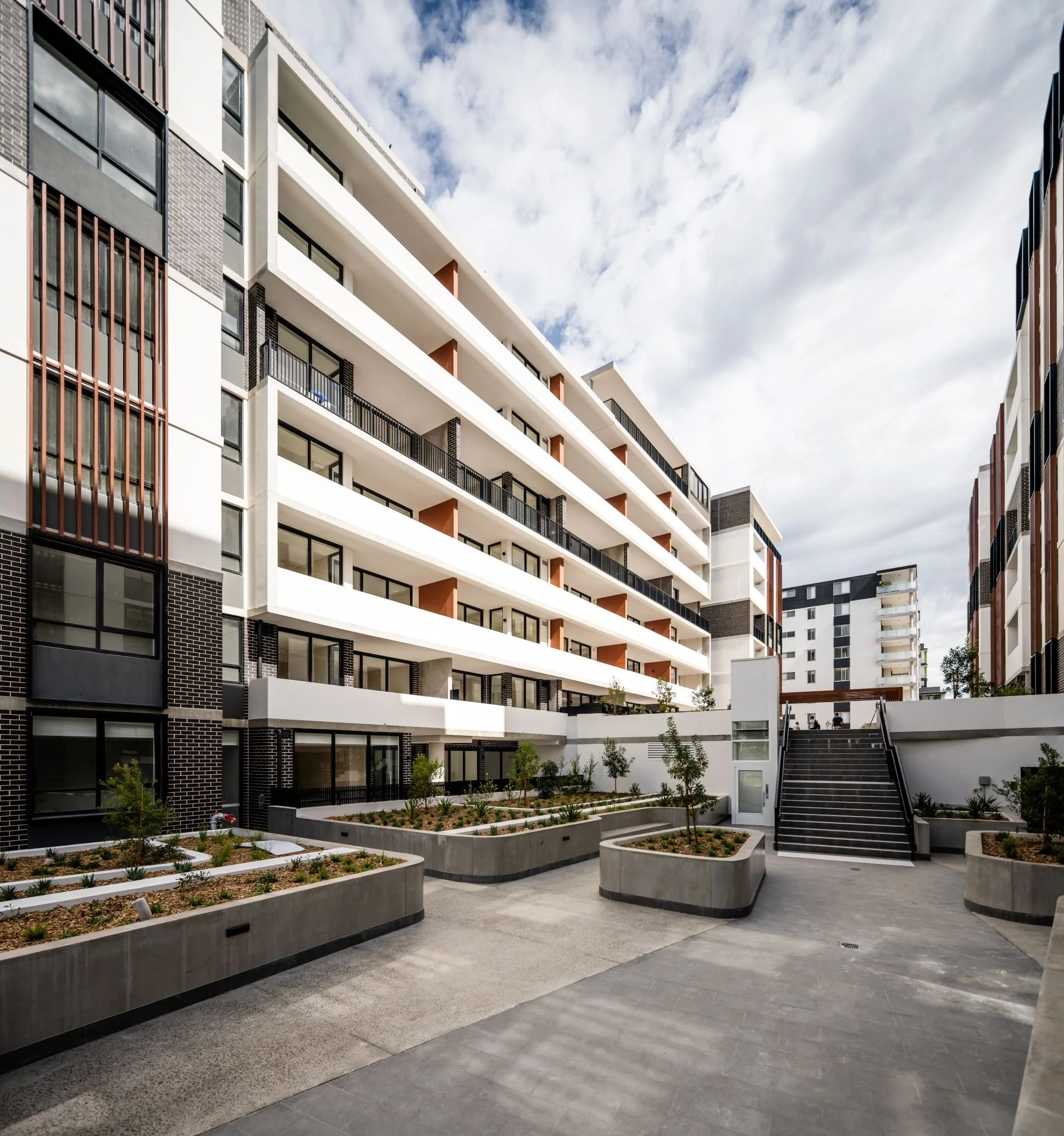 Modern multi-story apartment building with balconies, landscaped courtyard, and outdoor stairs, under a cloudy sky.