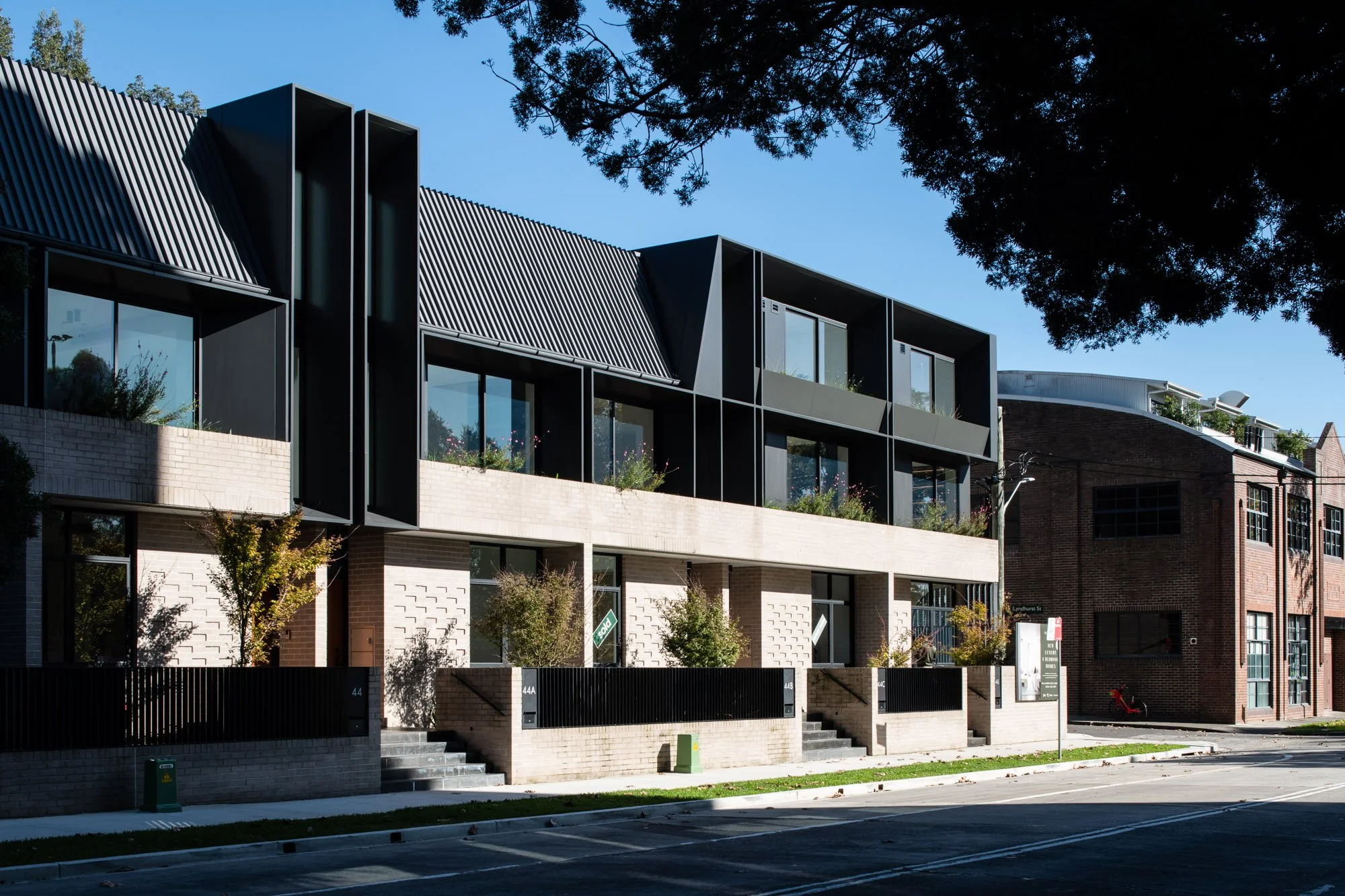 Modern multi-story residential building with black and beige exterior, large windows, and patio gardens on the balconies, situated on a street with trees and neighboring brick buildings.