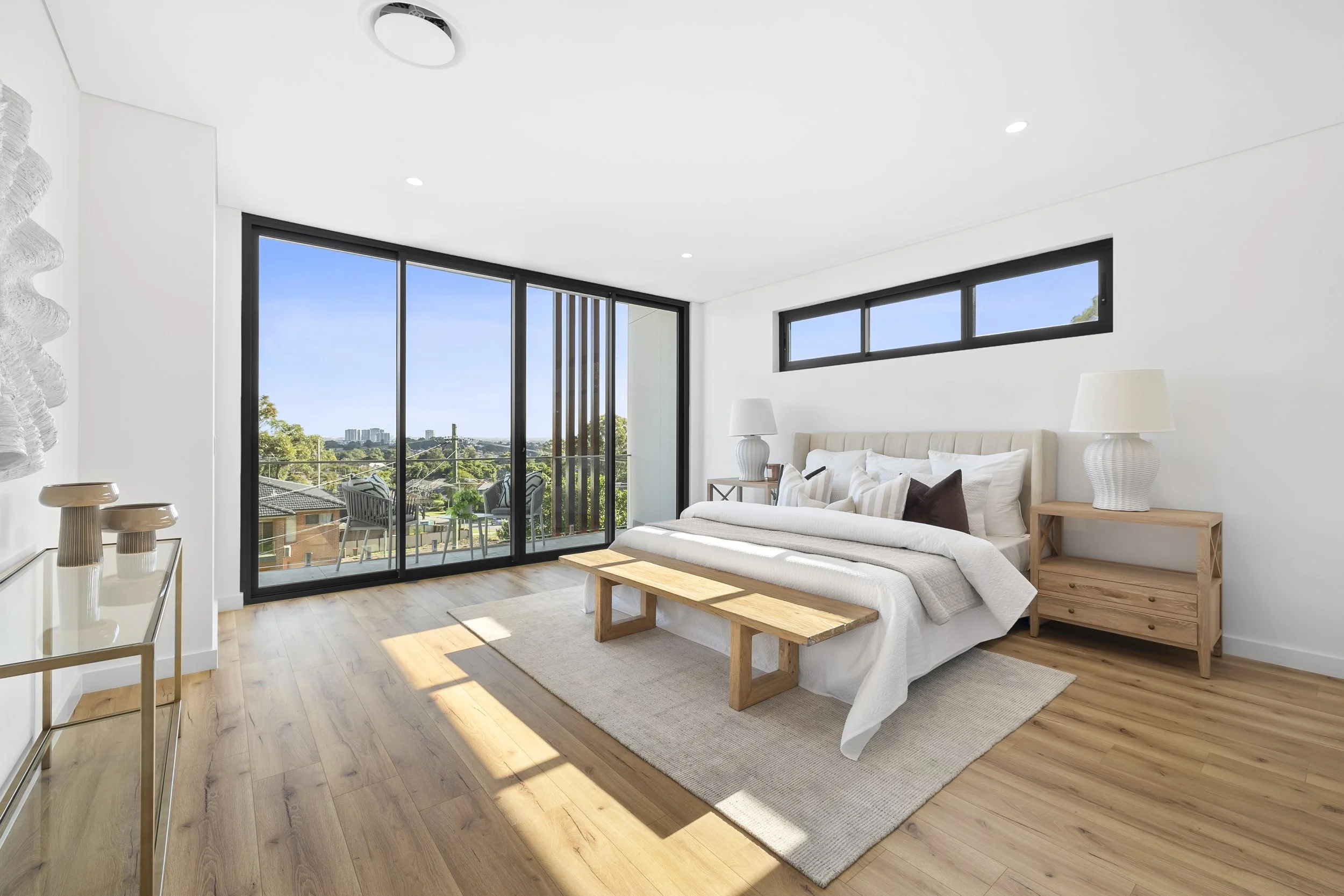 Modern bedroom with wooden floors, white walls, a large bed with beige headboard, white bedding, and multiple pillows, beside two wooden nightstands with matching lamps, a wooden bench at the foot of the bed, and a balcony with outdoor furniture seen