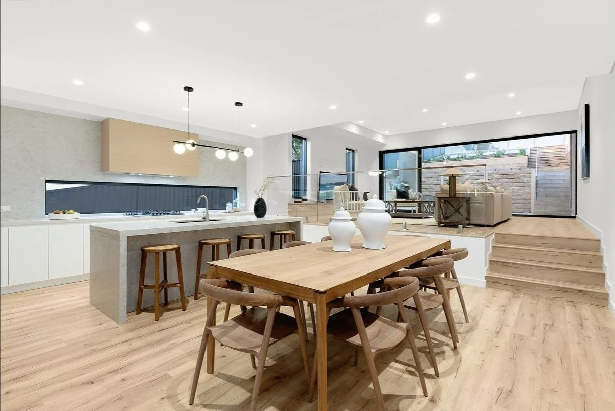 Open-concept kitchen and dining area with wooden flooring, modern light fixtures, a kitchen island with stools, a wooden dining table with chairs, and a sitting area with large glass sliding doors