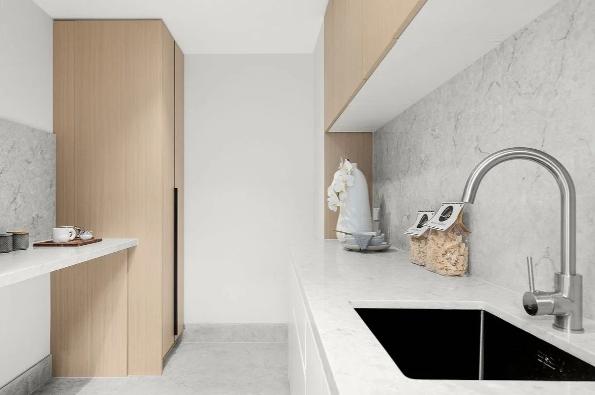 Modern kitchen with light wood cabinets, a white marble countertop, a black sink, and minimal decor including a vase with white flowers and jars of pasta.