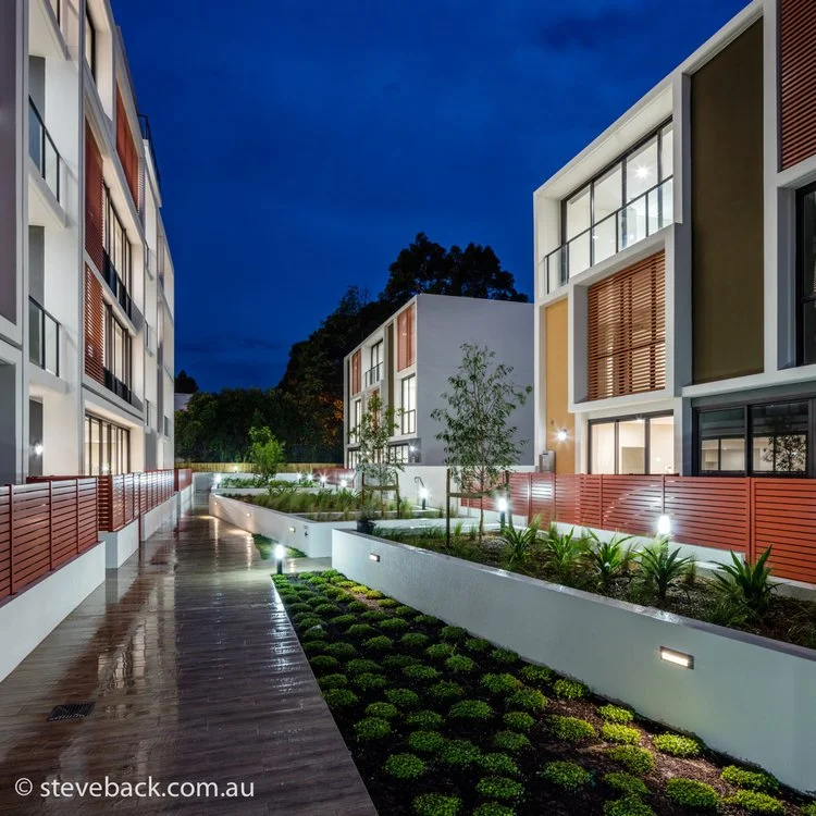 Modern residential complex with illuminated walkways, landscaped gardens, and contemporary multi-story buildings during twilight.
