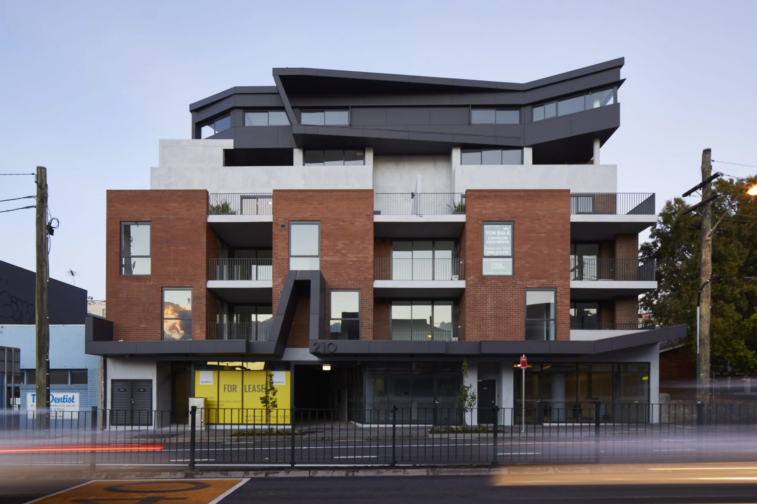 Modern multi-story building with brick and dark gray exterior, several balconies, large windows, and for sale and for lease signs, with a blurred vehicle in motion in the foreground.