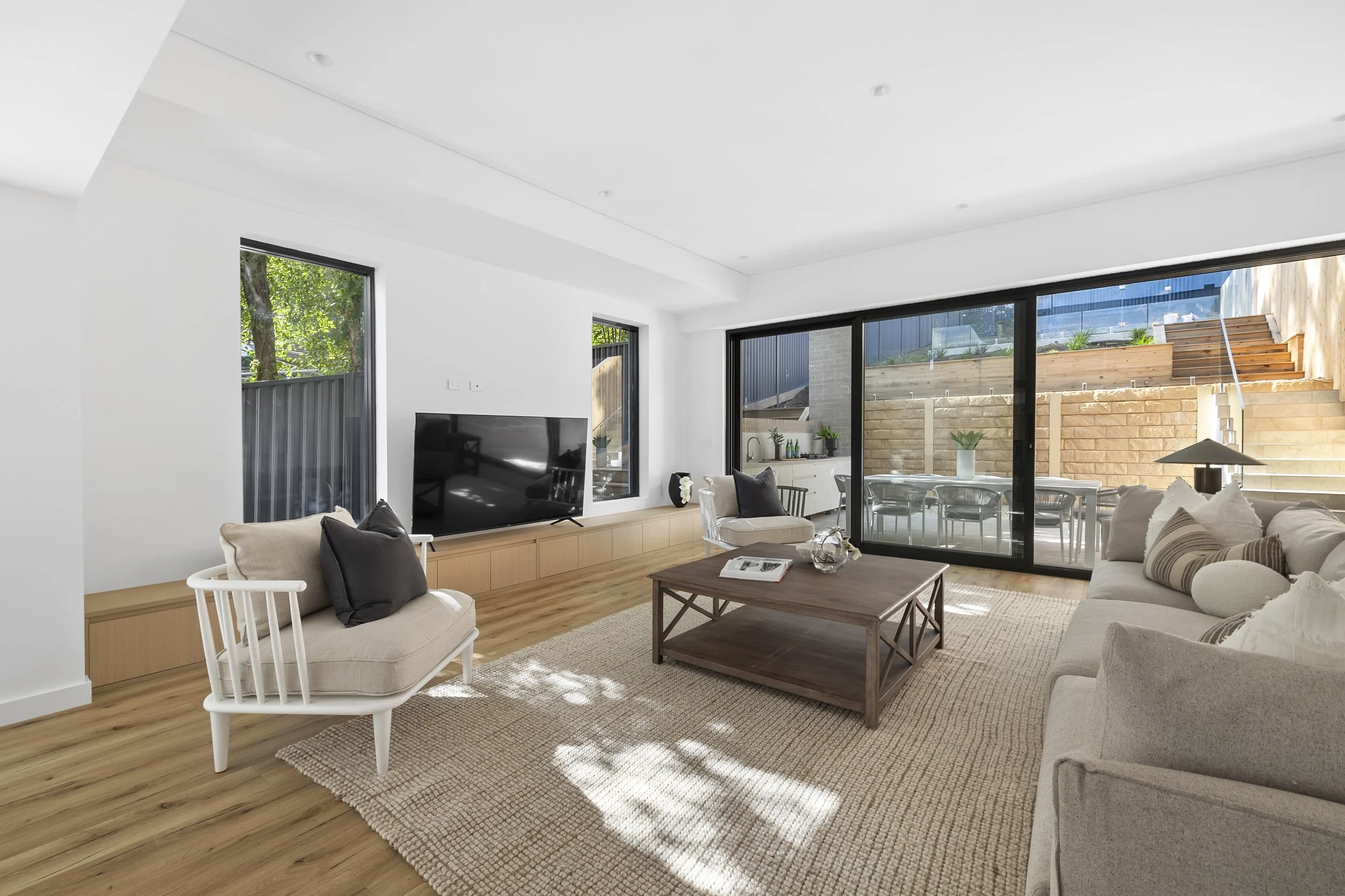 Modern living room with beige sofas, a wooden coffee table, a large flat-screen TV, hardwood flooring, and a sliding glass door leading to an outdoor patio with dining furniture and stairs.