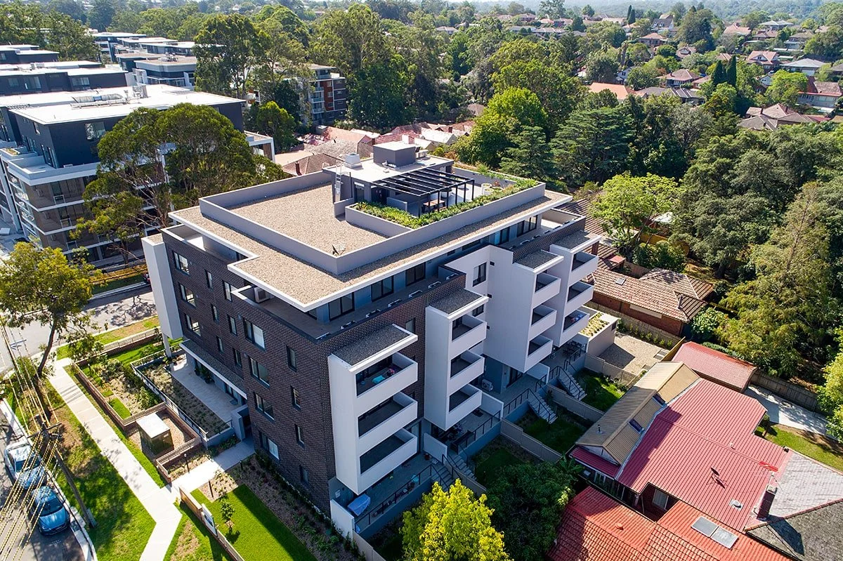 An aerial view of a modern, multi-story residential building with white and dark brick exterior walls, surrounded by greenery and neighboring houses, in a suburban neighborhood.