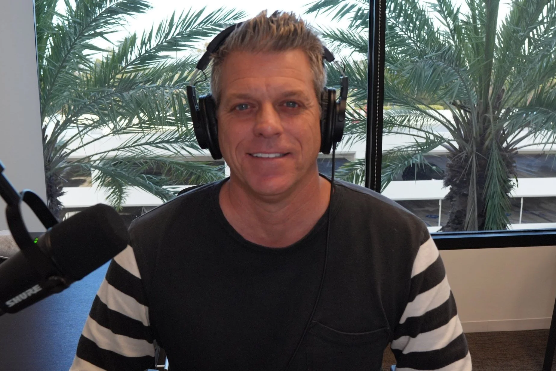 Man with headphones and microphone in office setting with palm trees outside the window.