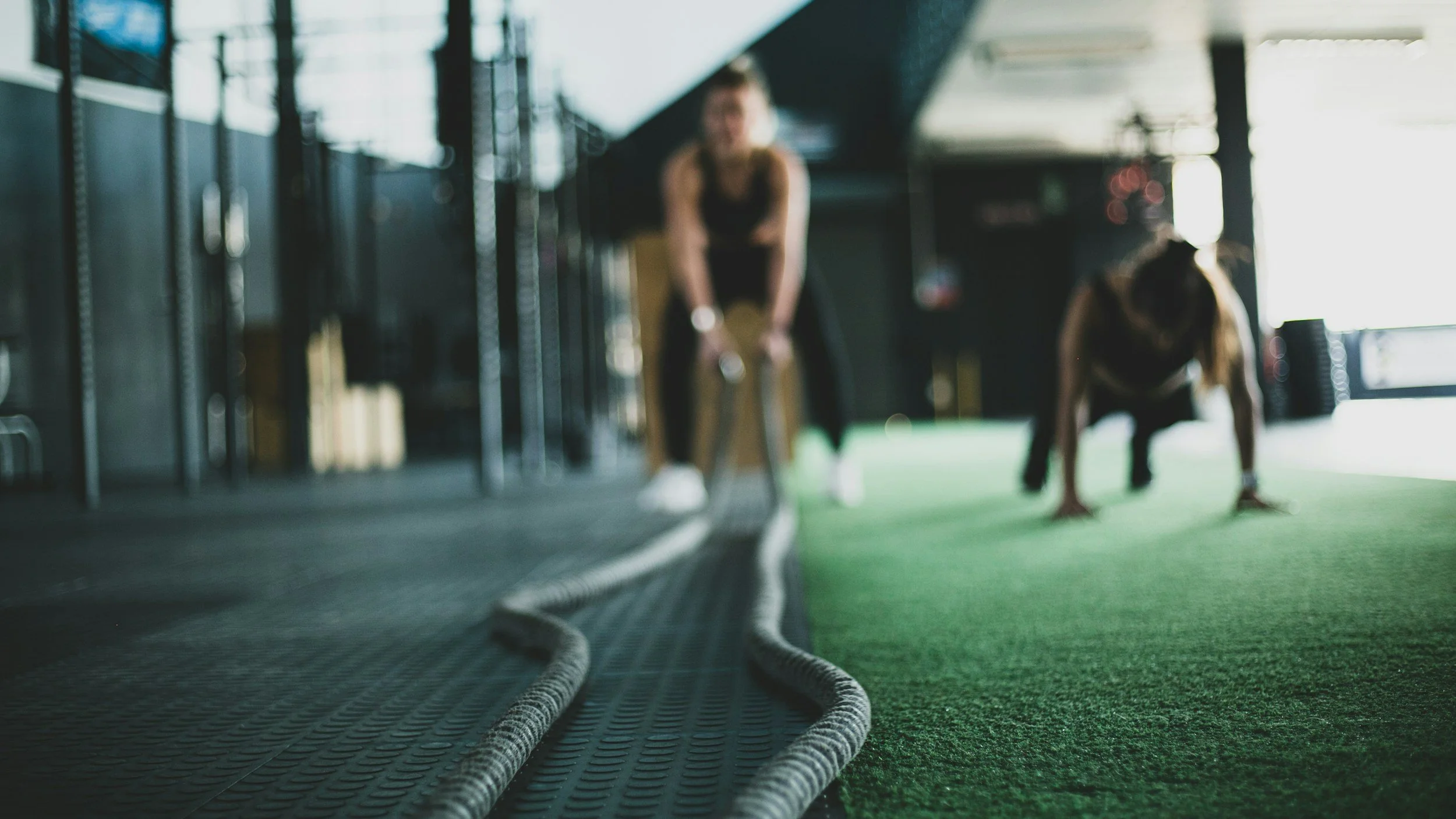 Two people exercising in a gym, using battle ropes, on a green turf and rubber floor.
