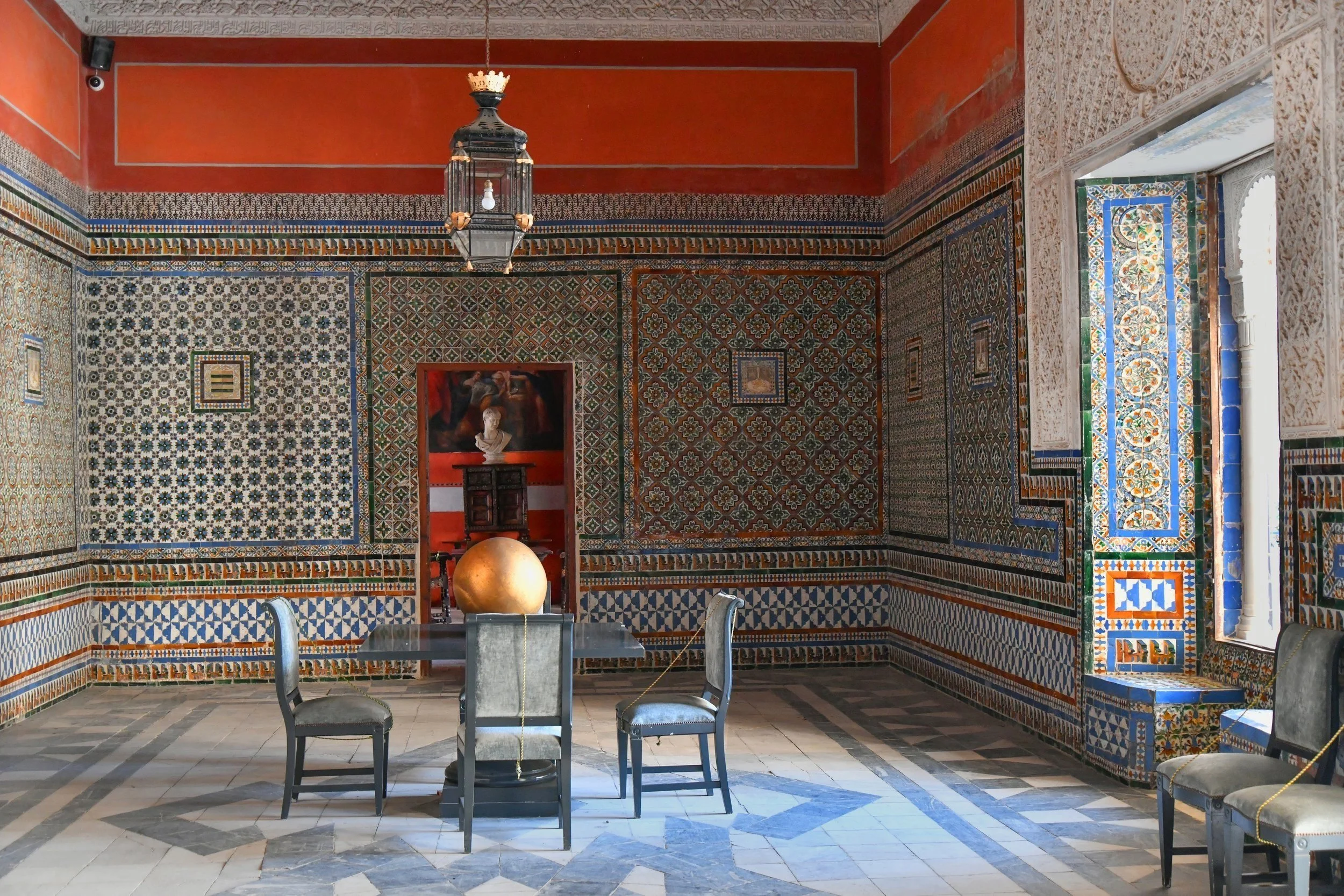 Decorative room with intricate tile patterns, featuring a central table with a golden globe, black chairs, and a hanging lamp. Walls are adorned with colorful geometric and floral tiles, while an open doorway reveals a bust sculpture in a red alcove.