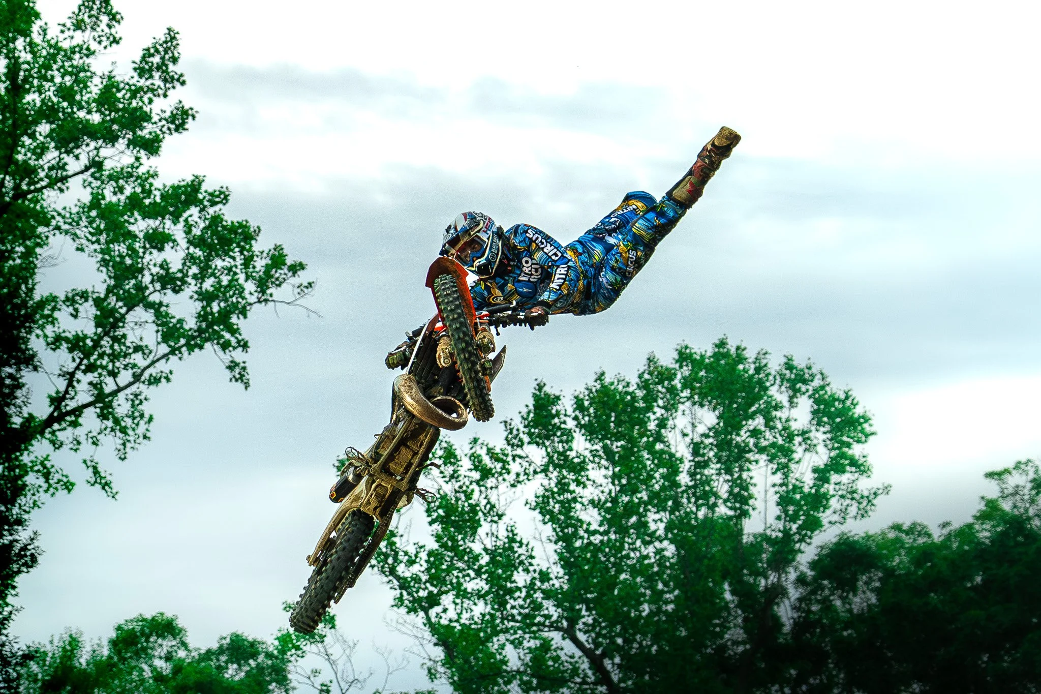 Motorcycle stunt rider airborne against cloudy sky, trees in background, performing a trick with body extended parallel to the ground.