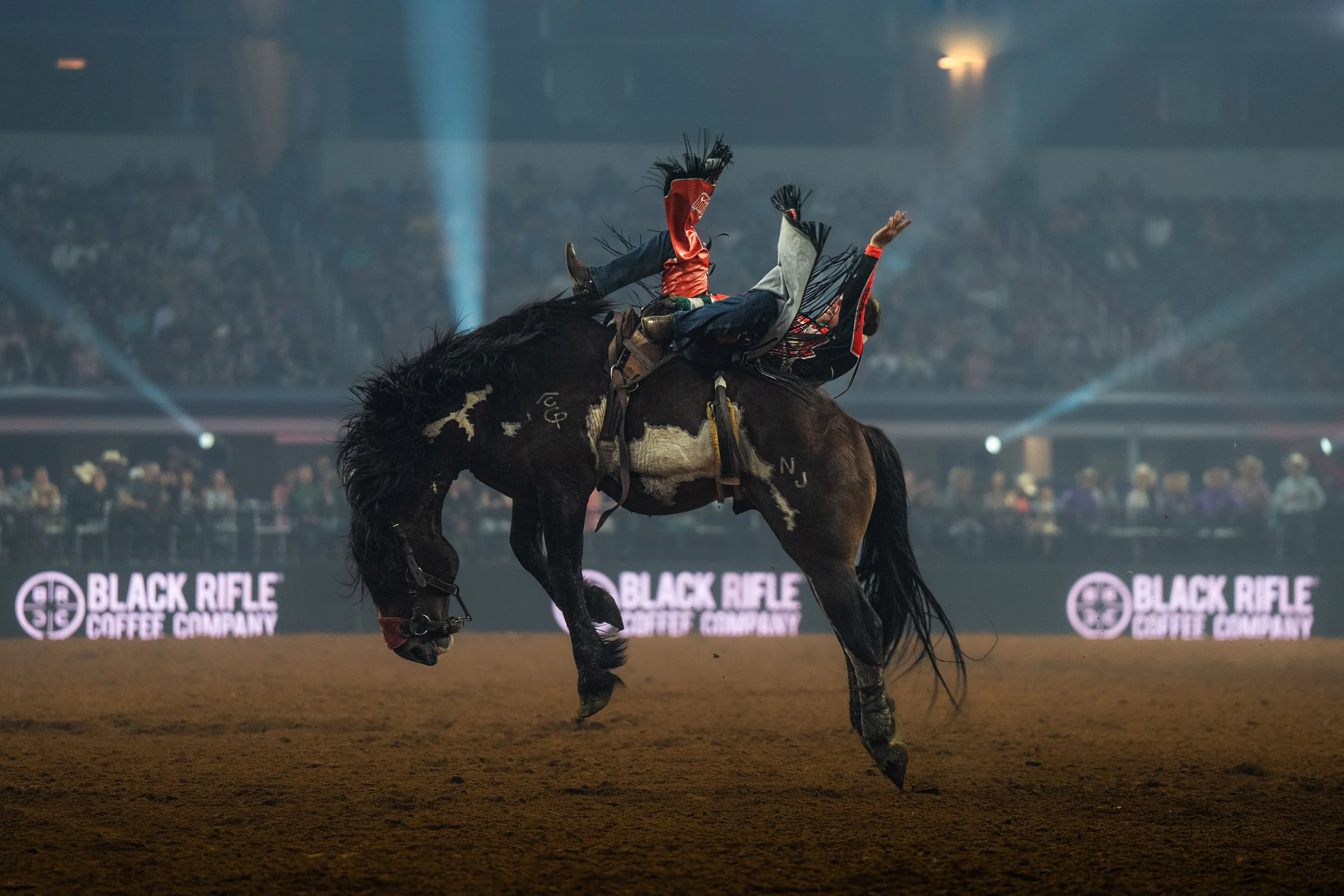 A rodeo rider falling off a bucking horse during a rodeo event at night, with a crowd in the background and branding for Black Rifle Coffee Company on the screens.