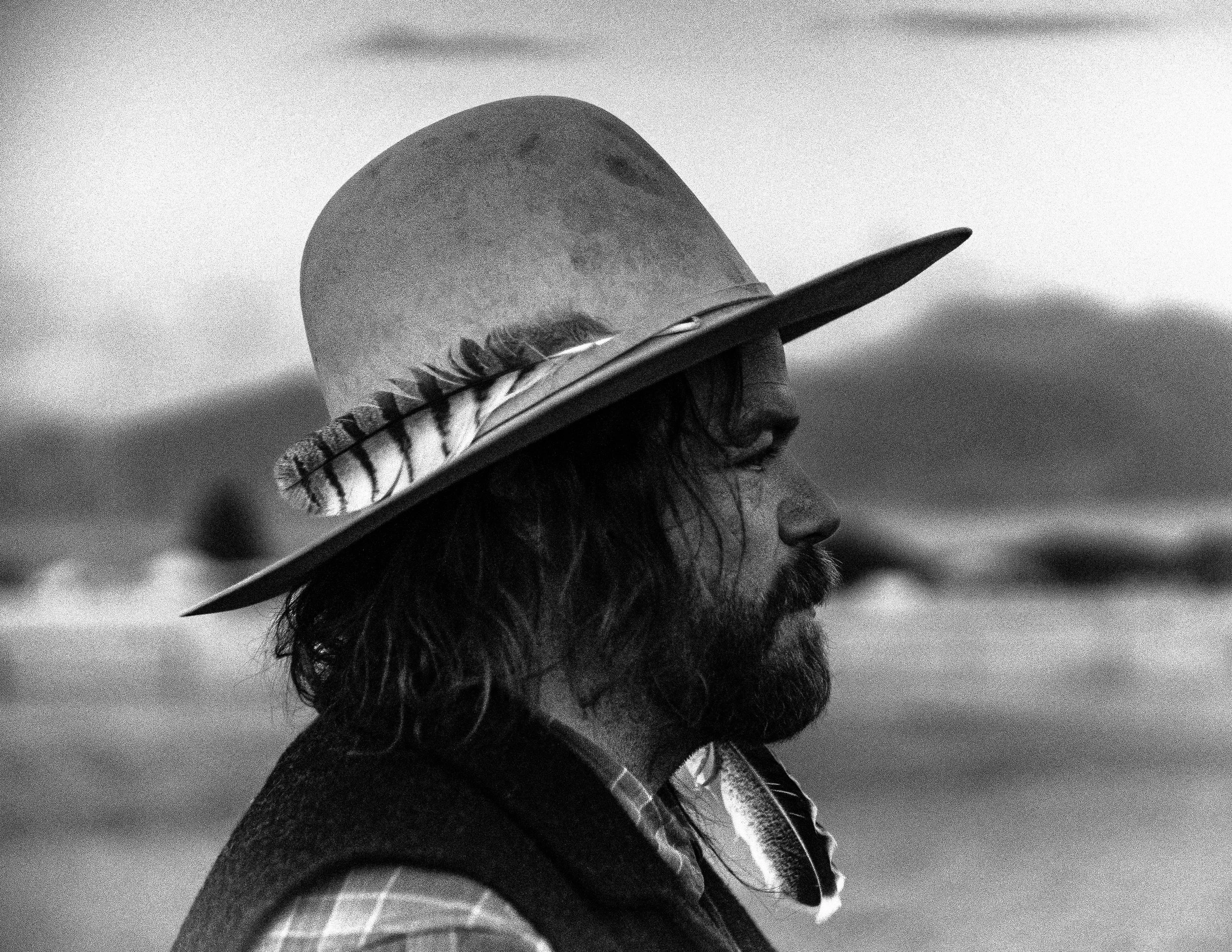 Side profile of a man with long hair and a beard wearing a wide-brimmed hat with feathers and a plaid shirt, outdoors with blurred landscape background.