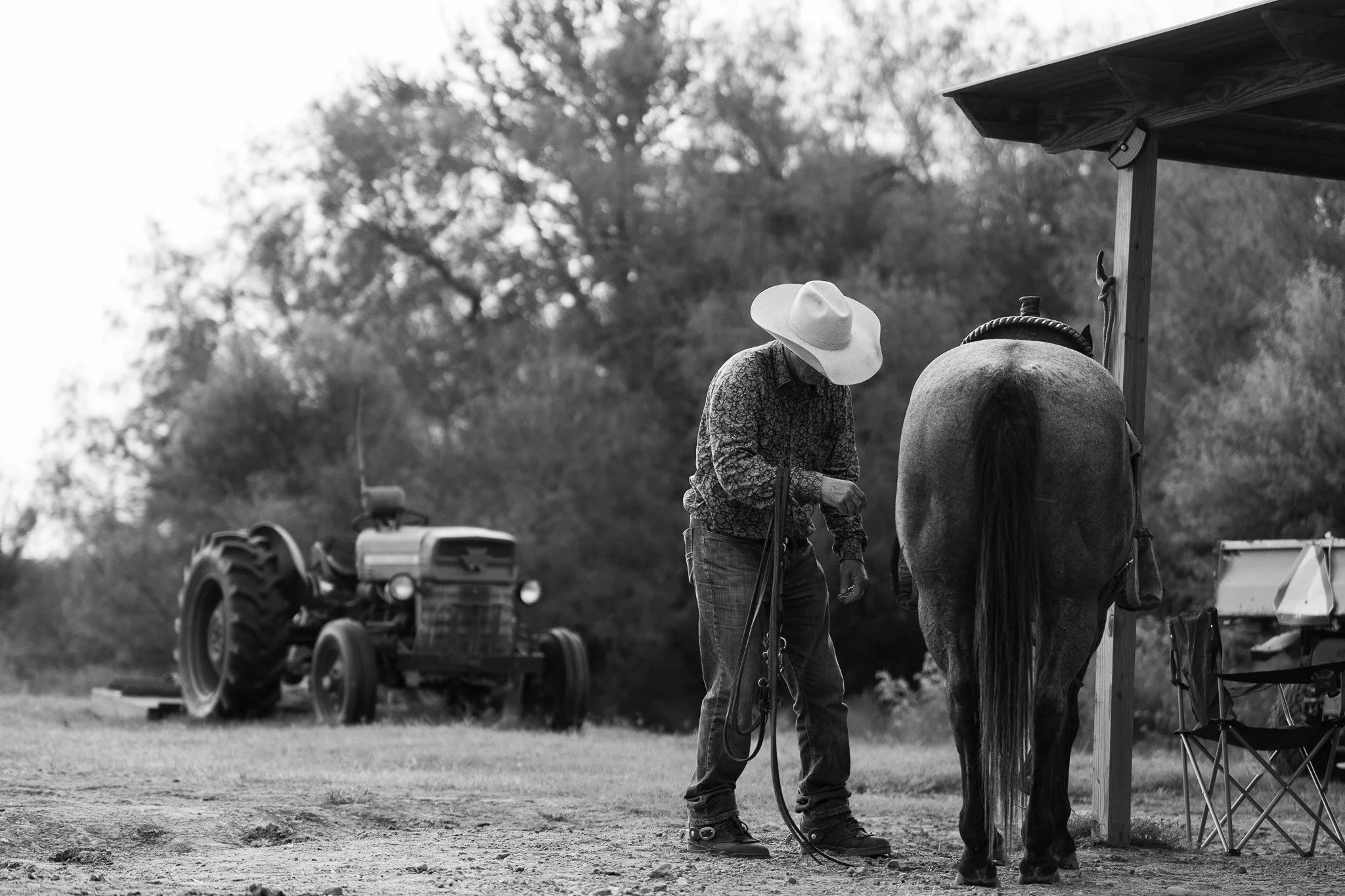 A person wearing a wide-brimmed hat standing next to a horse under a shelter, with a tractor in the background.