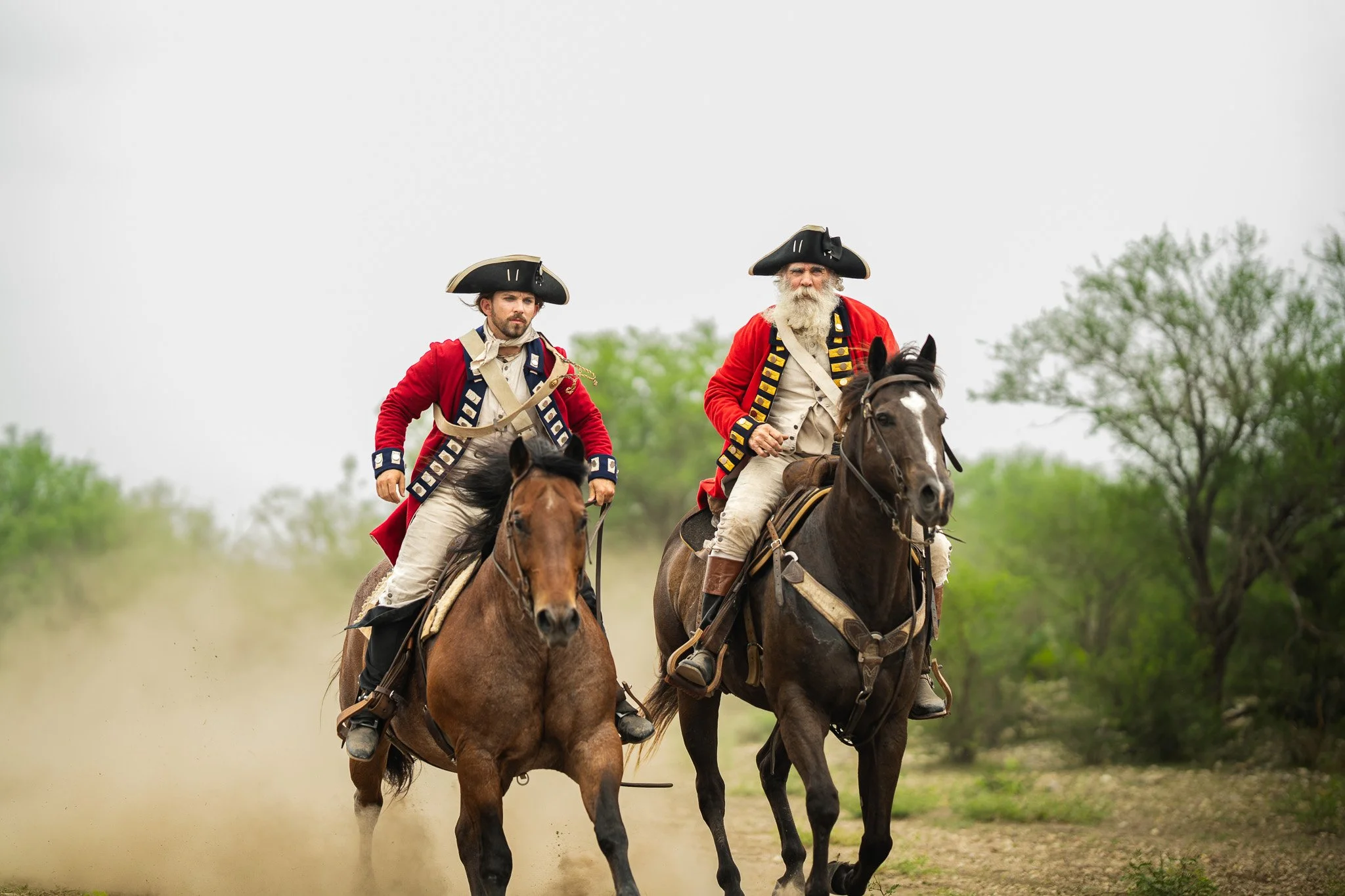 Two men dressed in Revolutionary War period military uniforms riding horses on a dirt trail with green trees in the background.