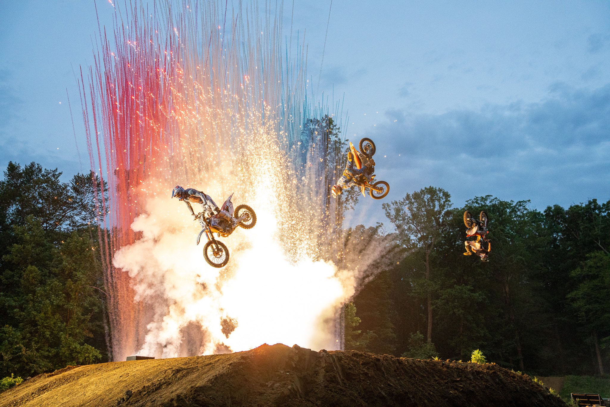 Four motocross riders performing stunts during a fireworks display at dusk, with trees in the background and a dirt track in the foreground.