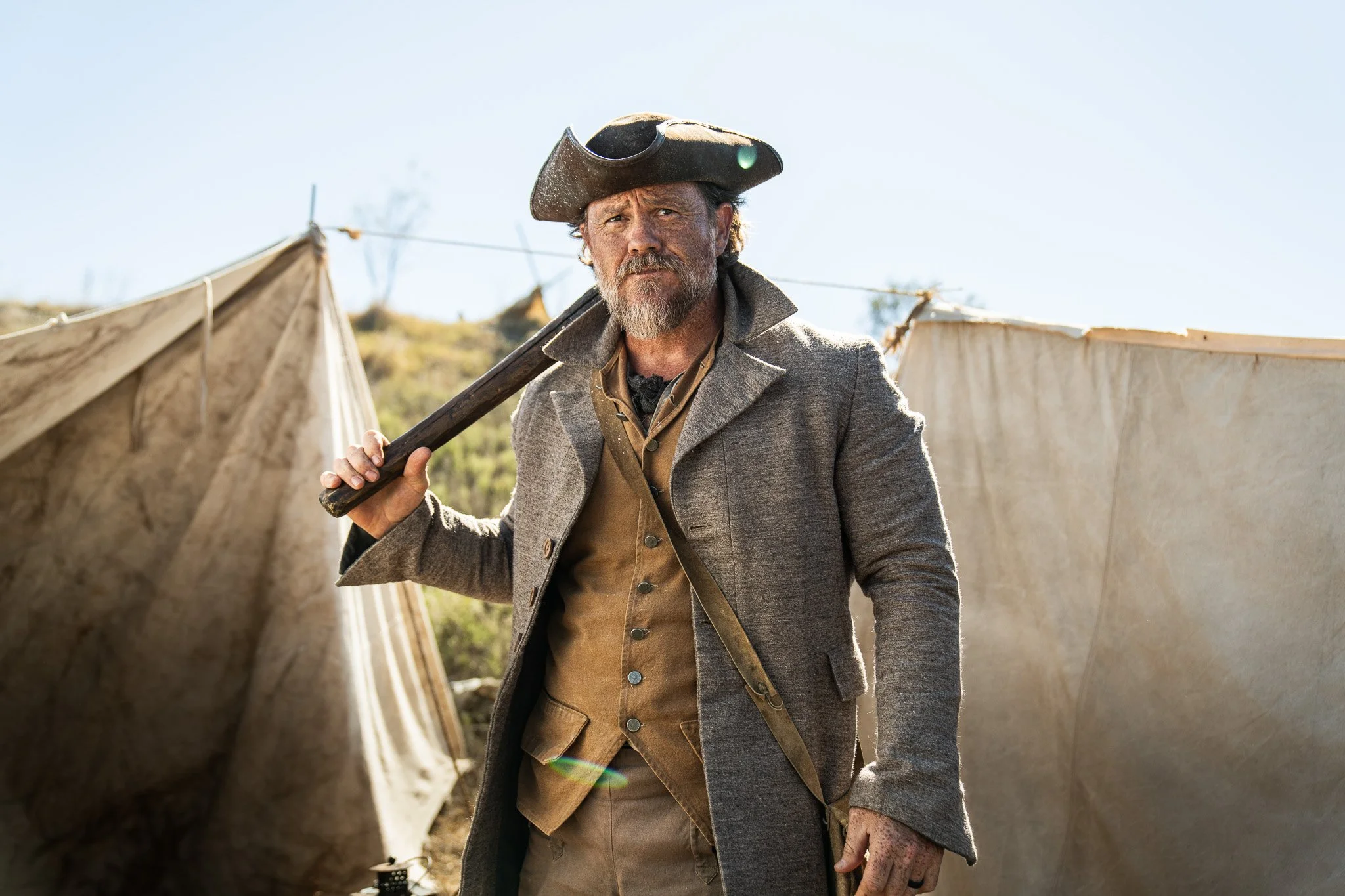 A man dressed as a historical settler or soldier in front of tents outdoors, holding a hatchet over his shoulder, wearing period clothing and a tricorn hat.