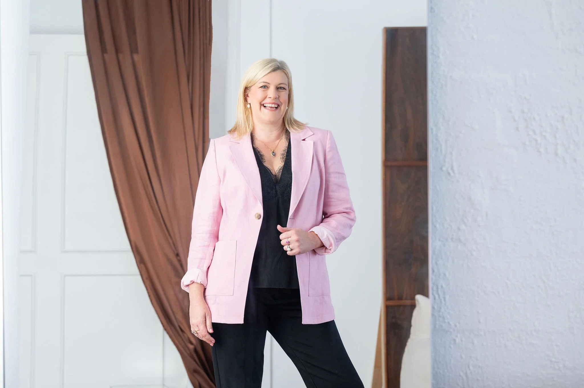 A woman with blonde hair, wearing a pink blazer and black outfit, smiling while standing indoors.