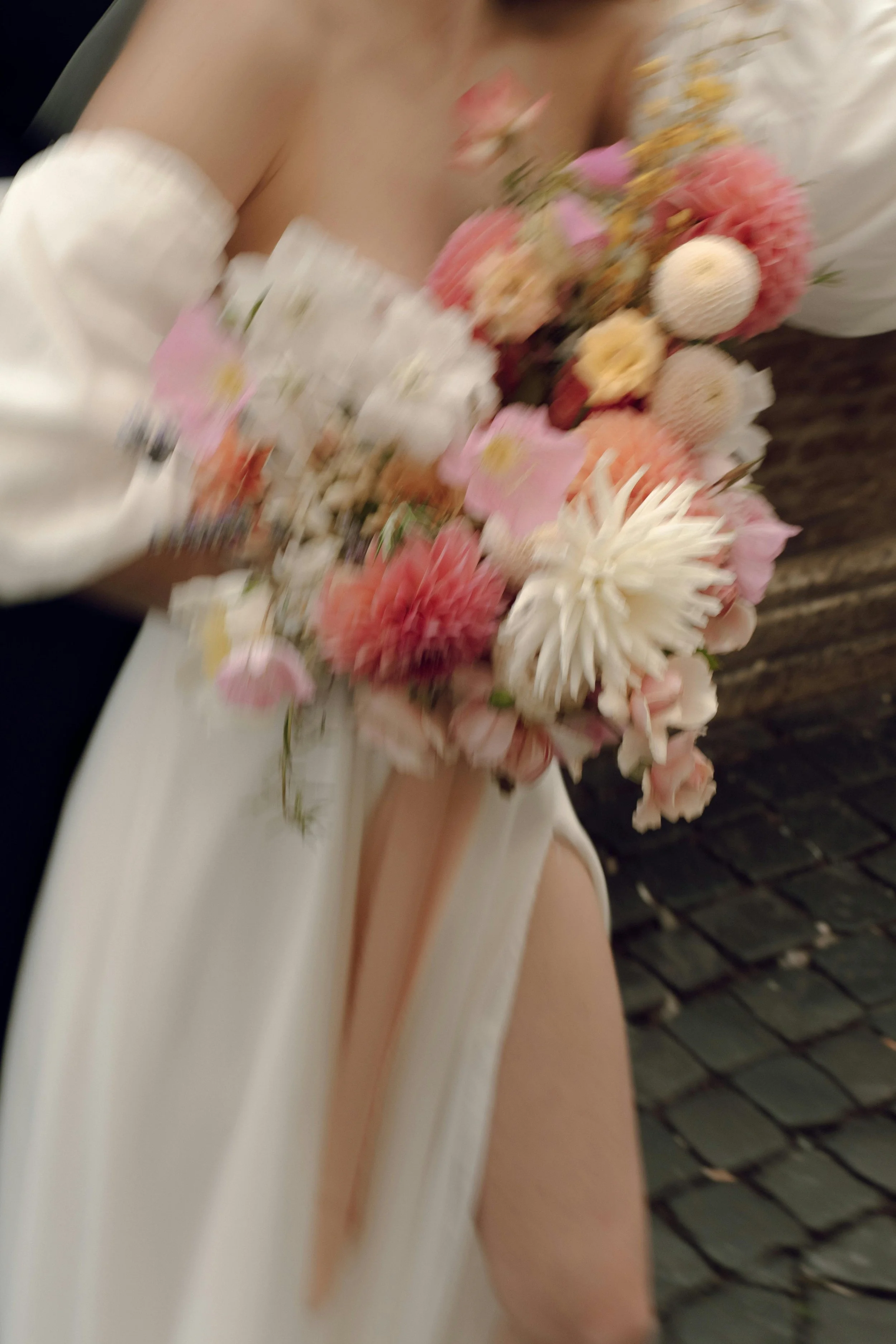 A person holding a bouquet of pink, white, and yellow flowers, standing next to a cobblestone wall.