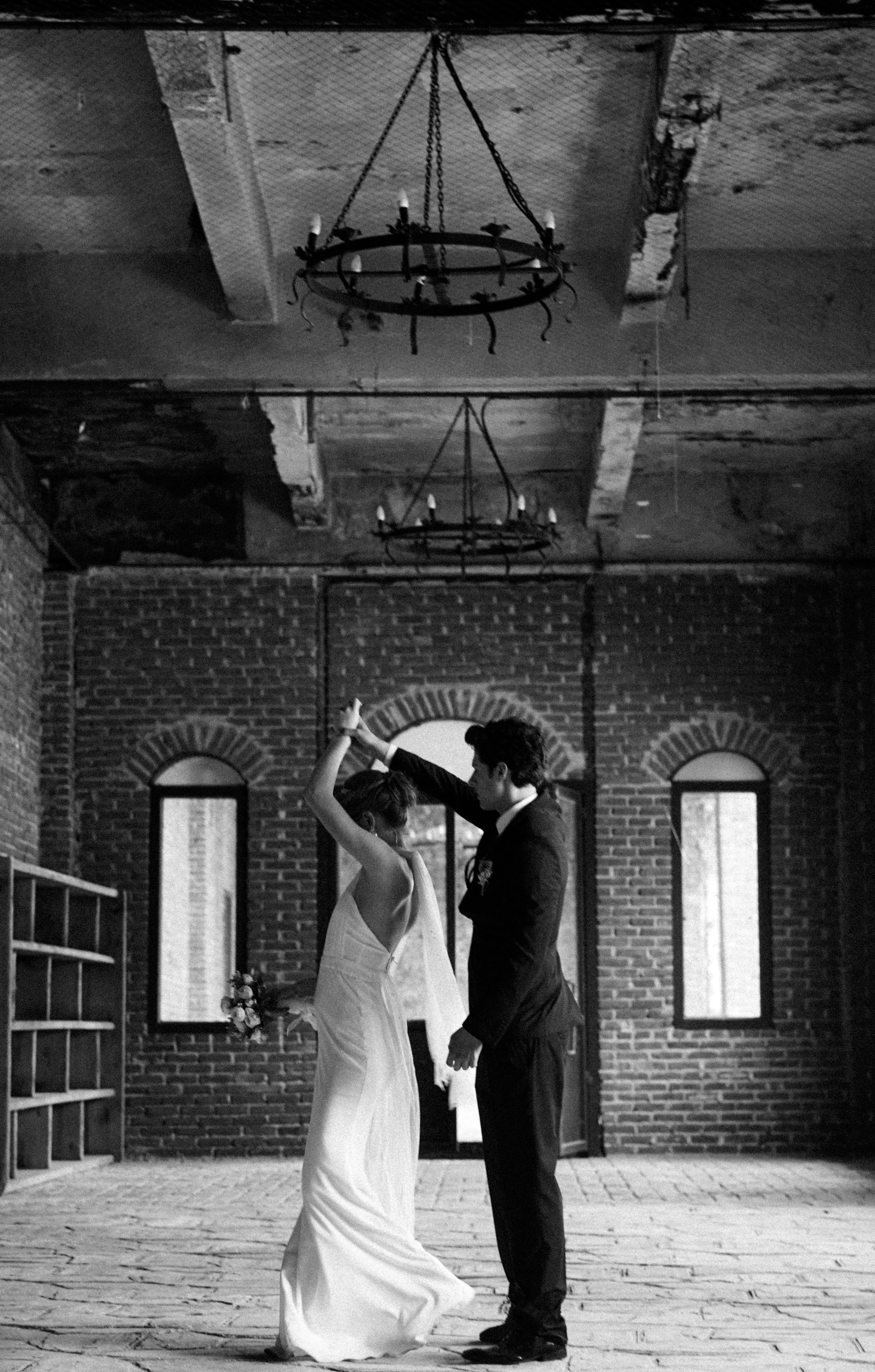 A black and white photo of a bride and groom dancing indoors. The bride wears a sleeveless wedding dress, and the groom is in a dark suit. They hold hands as they dance in front of a brick wall with three arched windows.