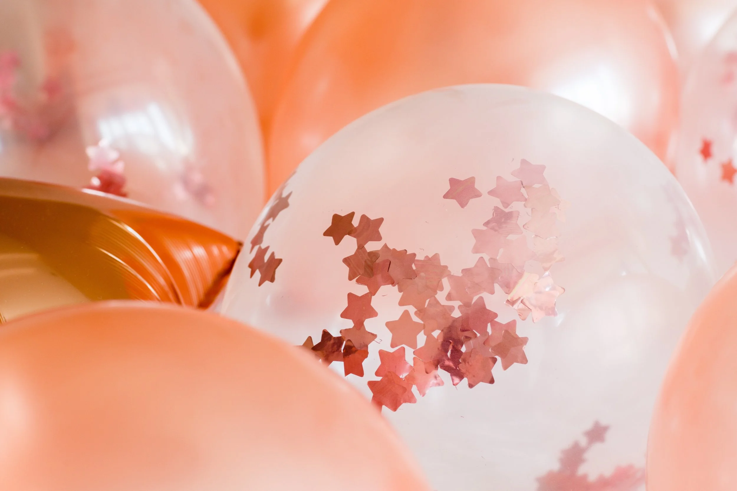 Close-up of pink, peach, and white balloons with a transparent balloon filled with pink star-shaped confetti.
