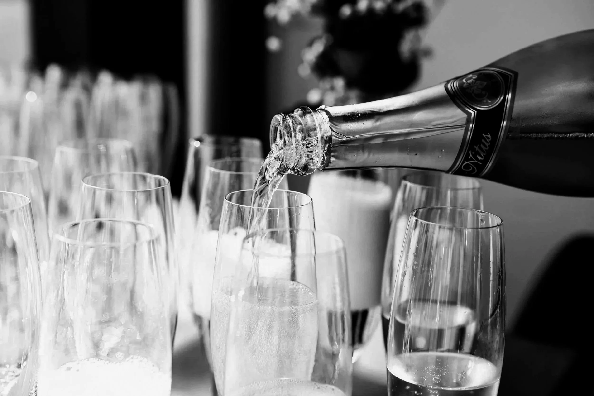 A bottle of champagne is being poured into several champagne flutes, with some already filled and others empty, on a table.