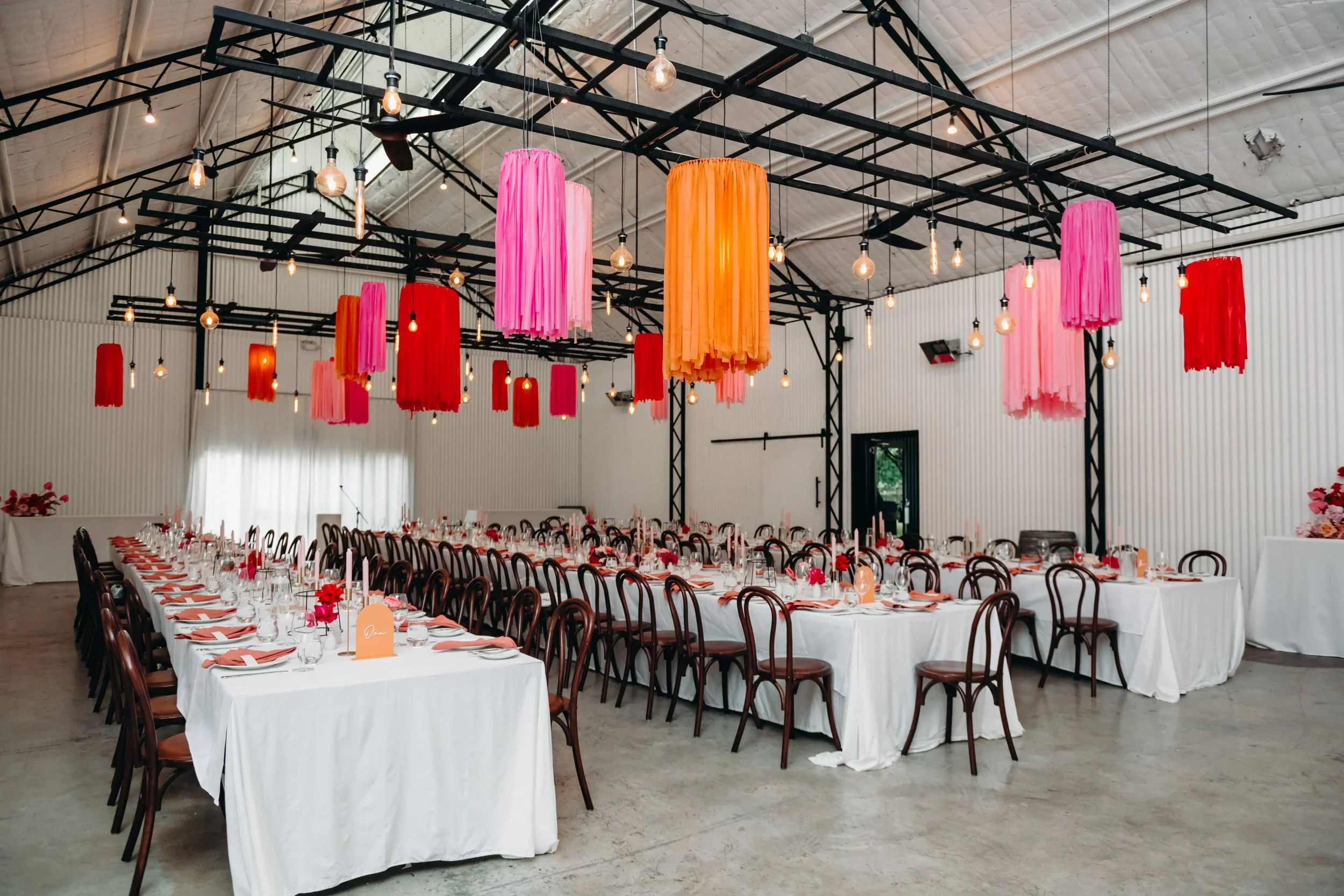 Event hall decorated with hanging pink, red, and orange paper lanterns, set with long tables covered in white tablecloths, arranged with glassware, cutlery, and floral centerpieces, ready for a celebration.