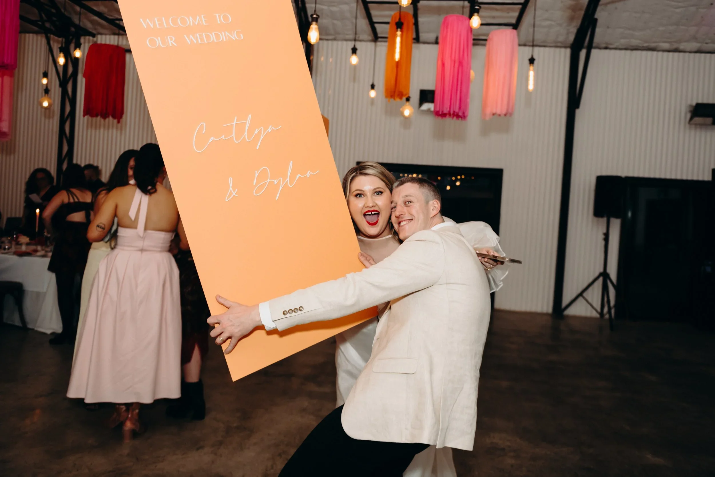 A couple hugging and smiling while holding a large orange wedding sign that reads 'Welcome to our wedding Caitlyn & Dylann' at a wedding reception with people, string lights, and hanging pink and orange decorations in the background.
