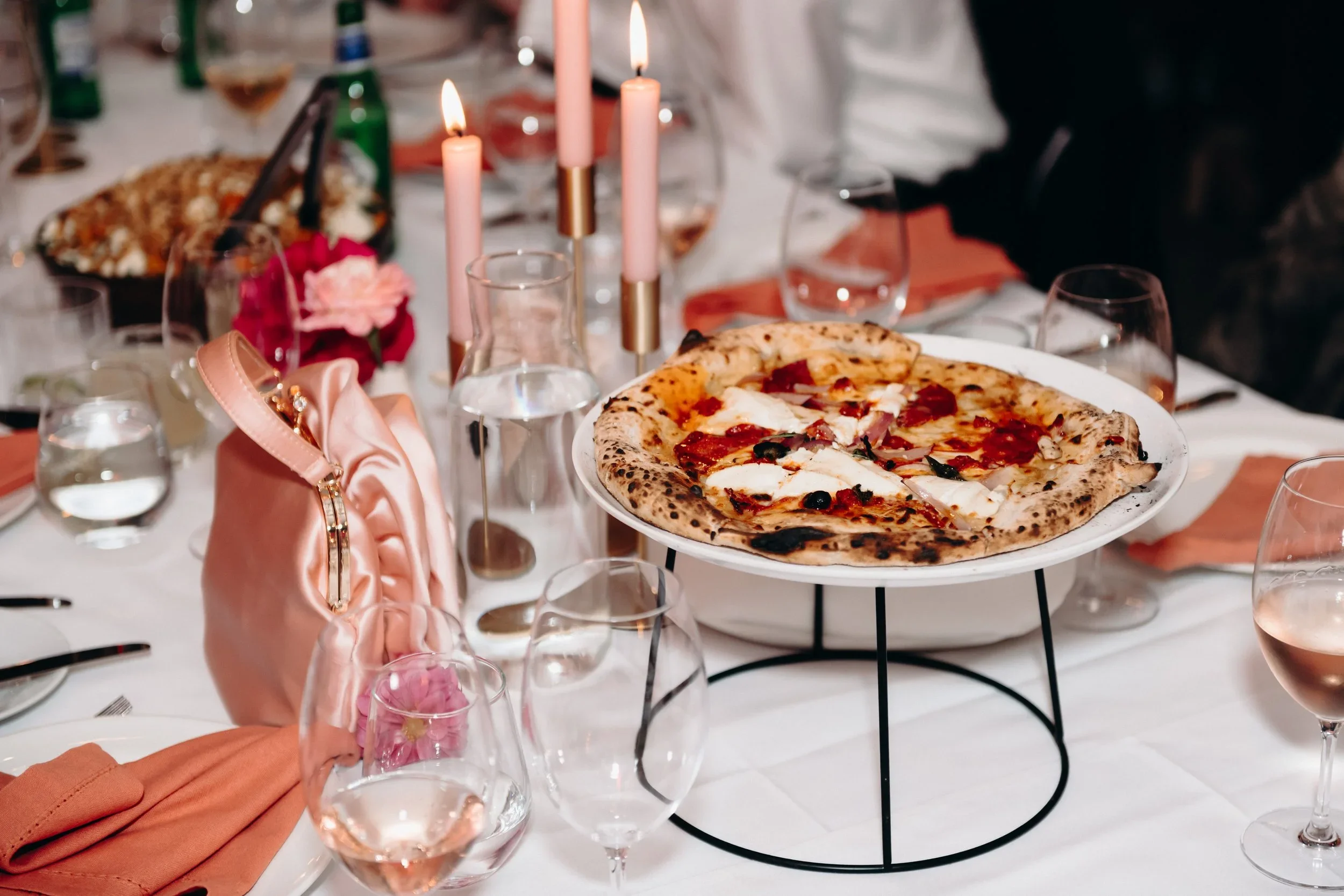 A table set for dining with a pizza on a stand, surrounded by glasses of water and wine, pink candles, a pink purse, and pink flowers, at a formal gathering.