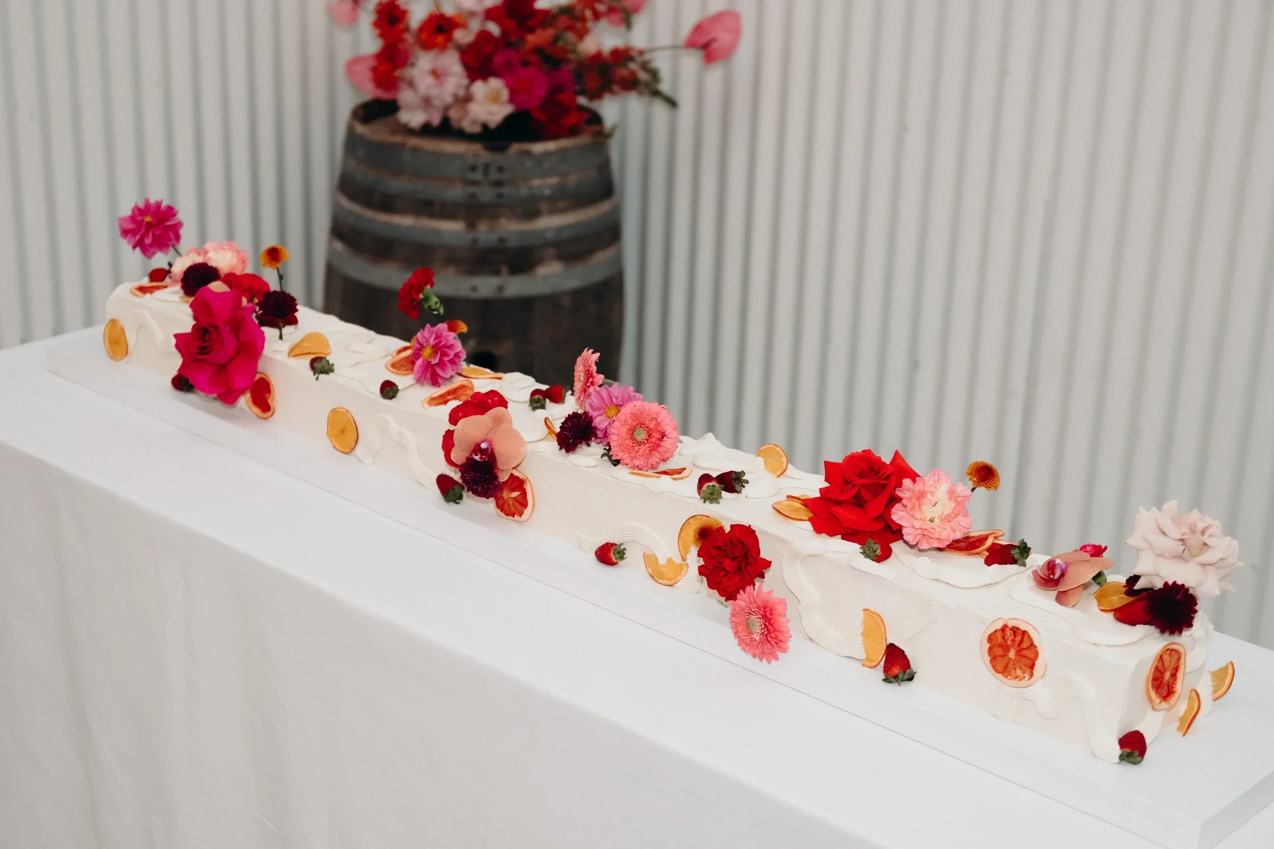 A long white wedding cake decorated with pink, red, and peach flowers, and dried orange slices, placed on a white tablecloth with a rustic barrel in the background.