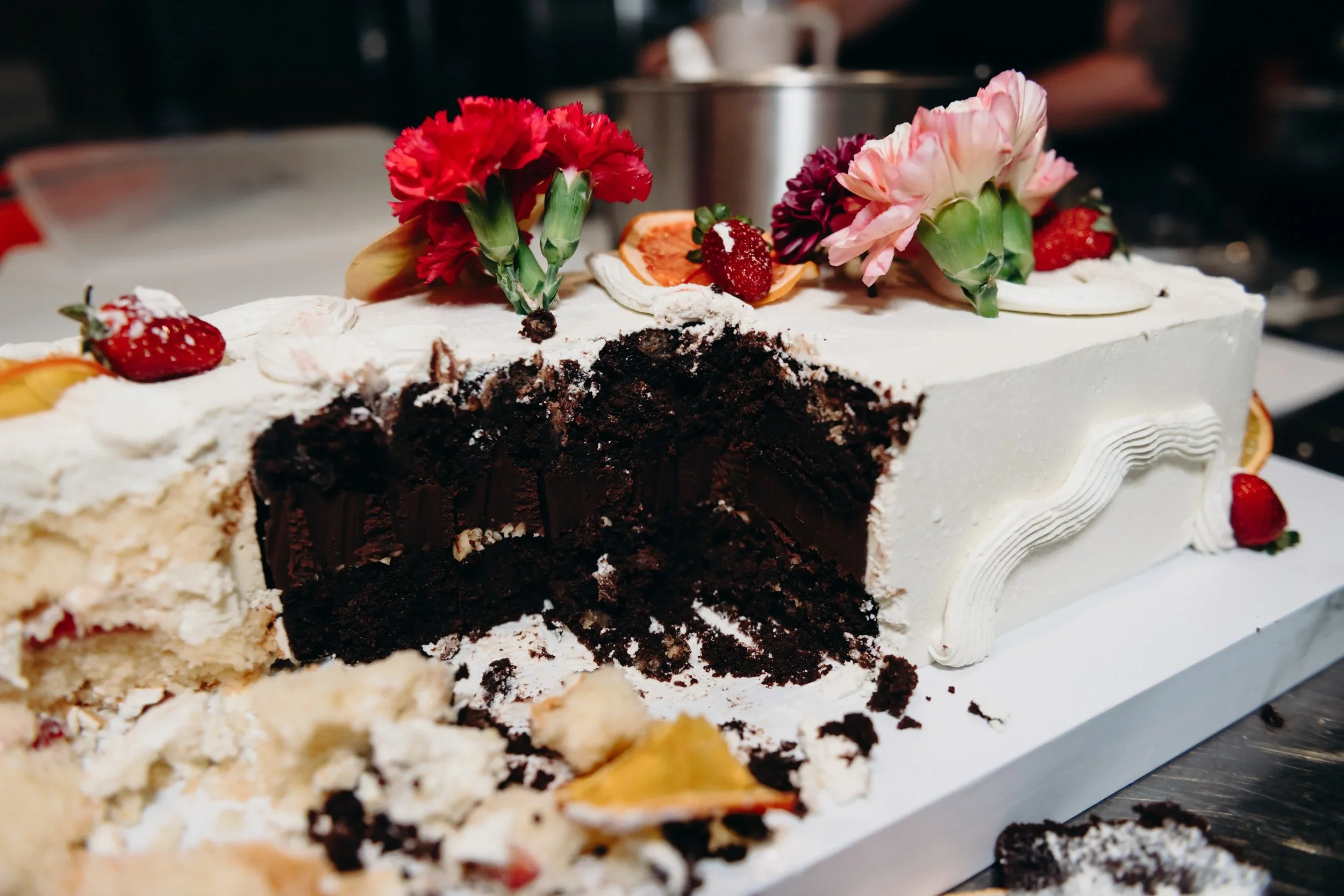 Chocolate layer cake with white frosting, decorated with strawberries, flowers, and slices of orange on top, with a portion missing.