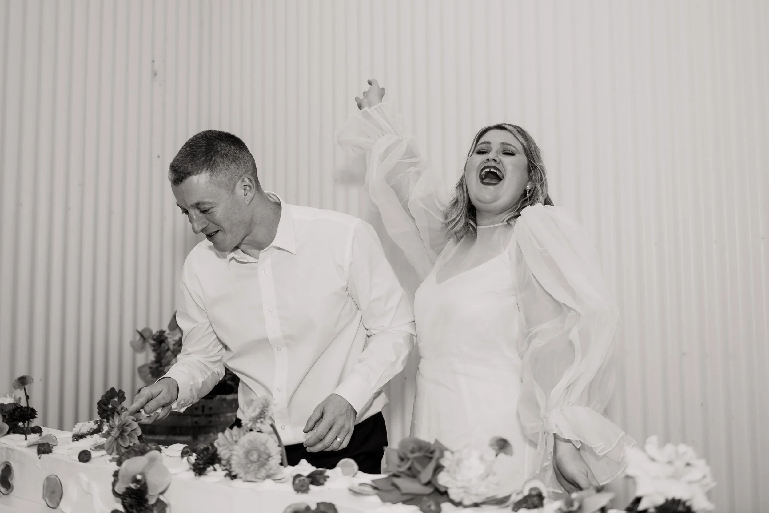 A black and white photo of a happy couple celebrating at a wedding reception. The woman is wearing a dress with puffed sleeves and has her arm raised, smiling widely. The man is wearing a white shirt and is focused on cutting a cake decorated with flowers.