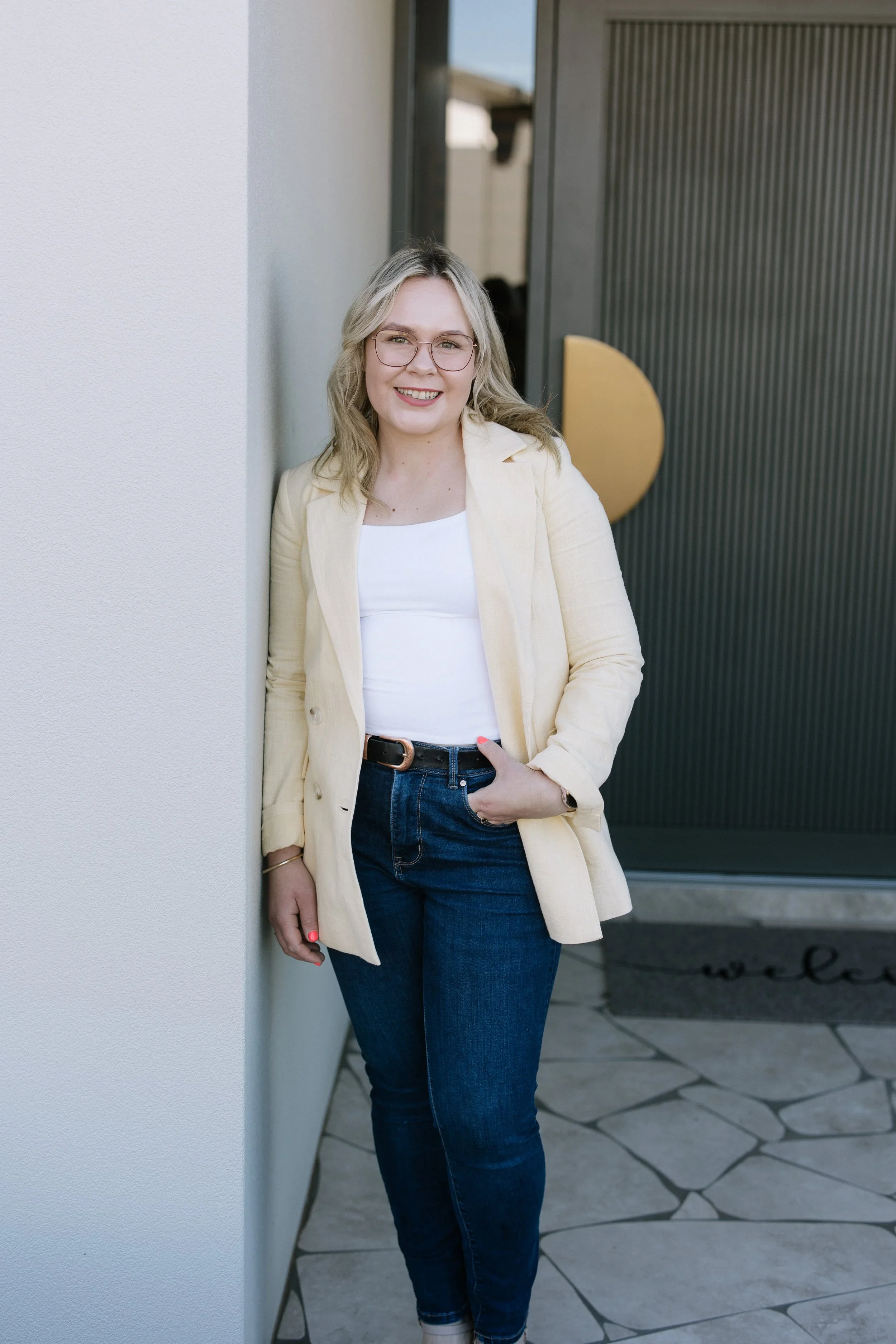 A young woman with blonde hair and glasses smiling while standing outdoors next to a white wall, wearing a cream blazer, white top, and dark jeans.